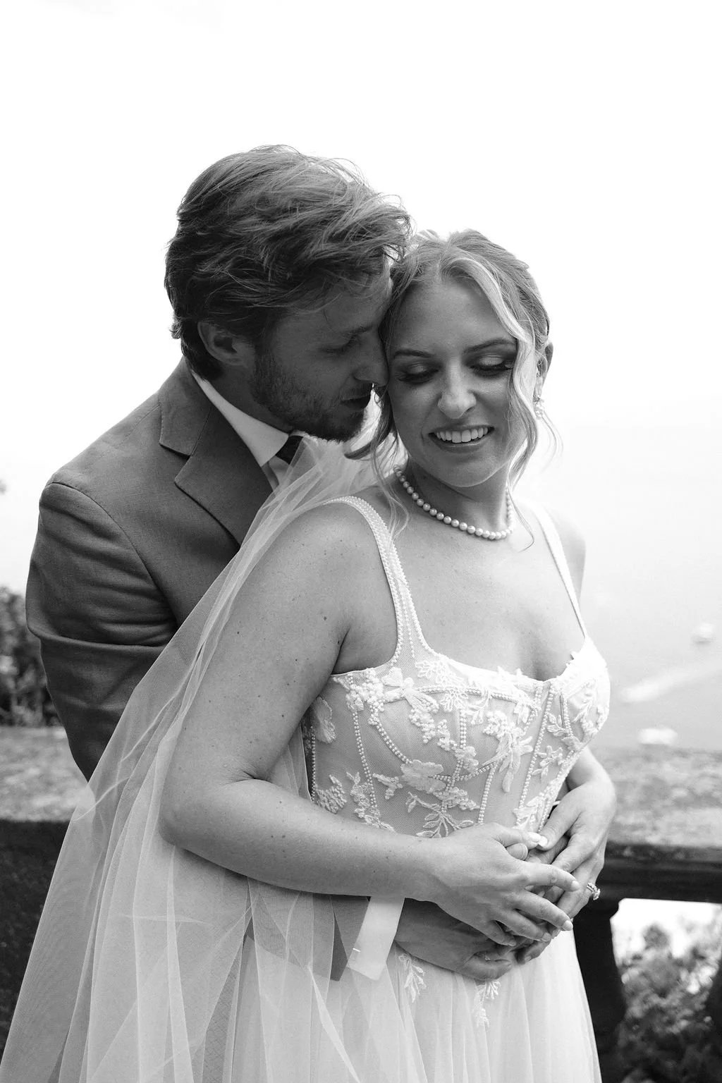 Black and white photo of a bride and groom close together, the groom whispering or leaning into the bride who is smiling, outdoors with a blurred background.