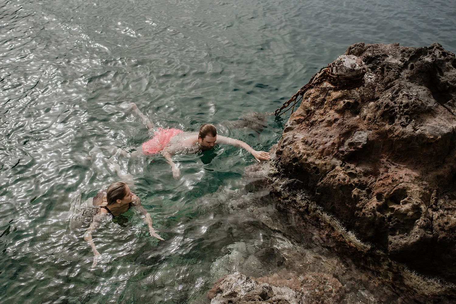 Two women swimming in the ocean near a large rock with chains attached to it.