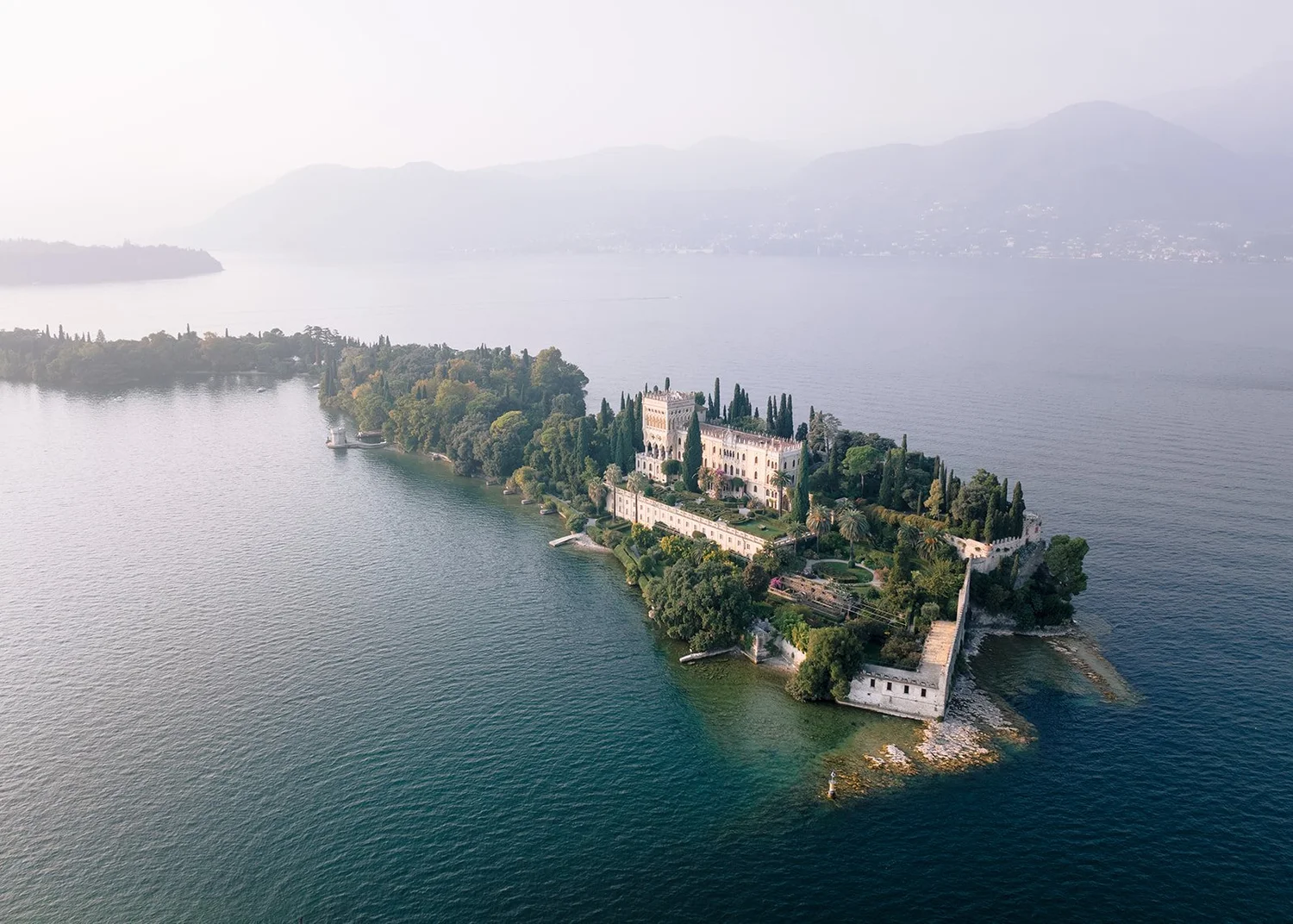 Aerial view of an island with a historic castle surrounded by dense trees and water, with mountains in the background.
