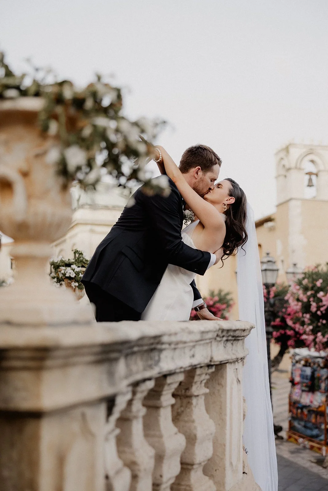 A bride and groom sharing a kiss on a balcony during their wedding, with flowers and historical architecture in the background.