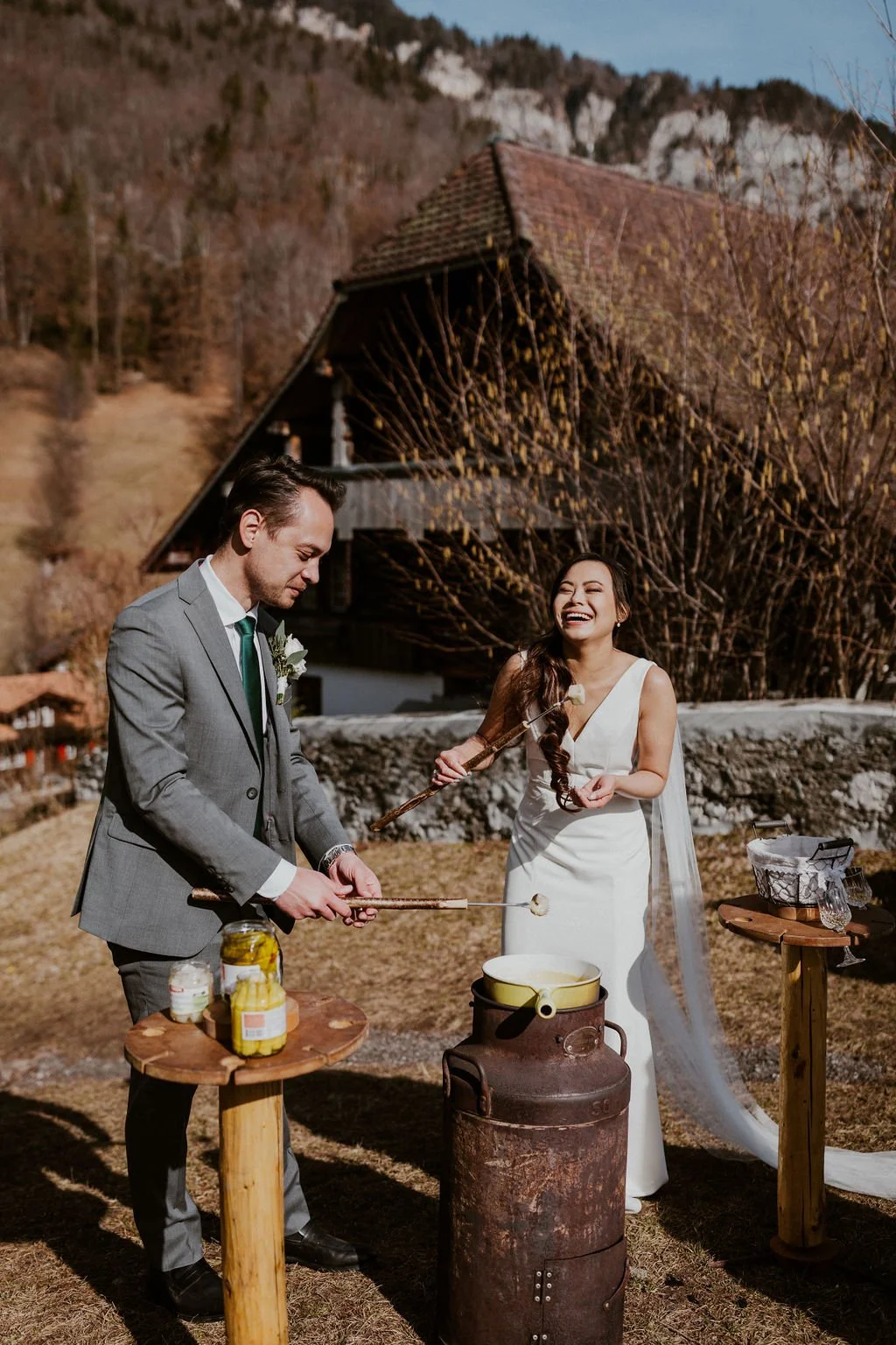 A bride and groom celebrating outdoors, cooking food over an open fire with wooden sticks, surrounded by a rustic setting with a house and mountains in the background, during daytime.