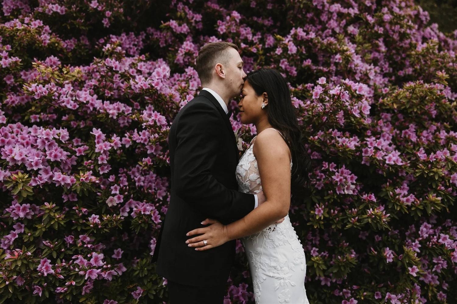 A newlywed couple stands close together with their foreheads touching, in front of a backdrop of pink azalea flowers. The groom is dressed in a black tuxedo and the bride in a white lace wedding gown, both appearing tender and emotional.