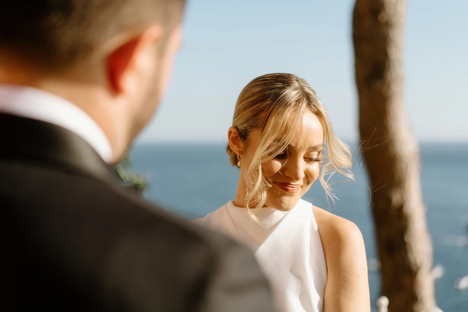 A smiling woman with blonde hair styled in an elegant updo, dressed in a white sleeveless dress, is photographed outdoors with a blurred ocean and a tree trunk in the background. A man in a dark suit is partially visible, slightly out of focus, in th