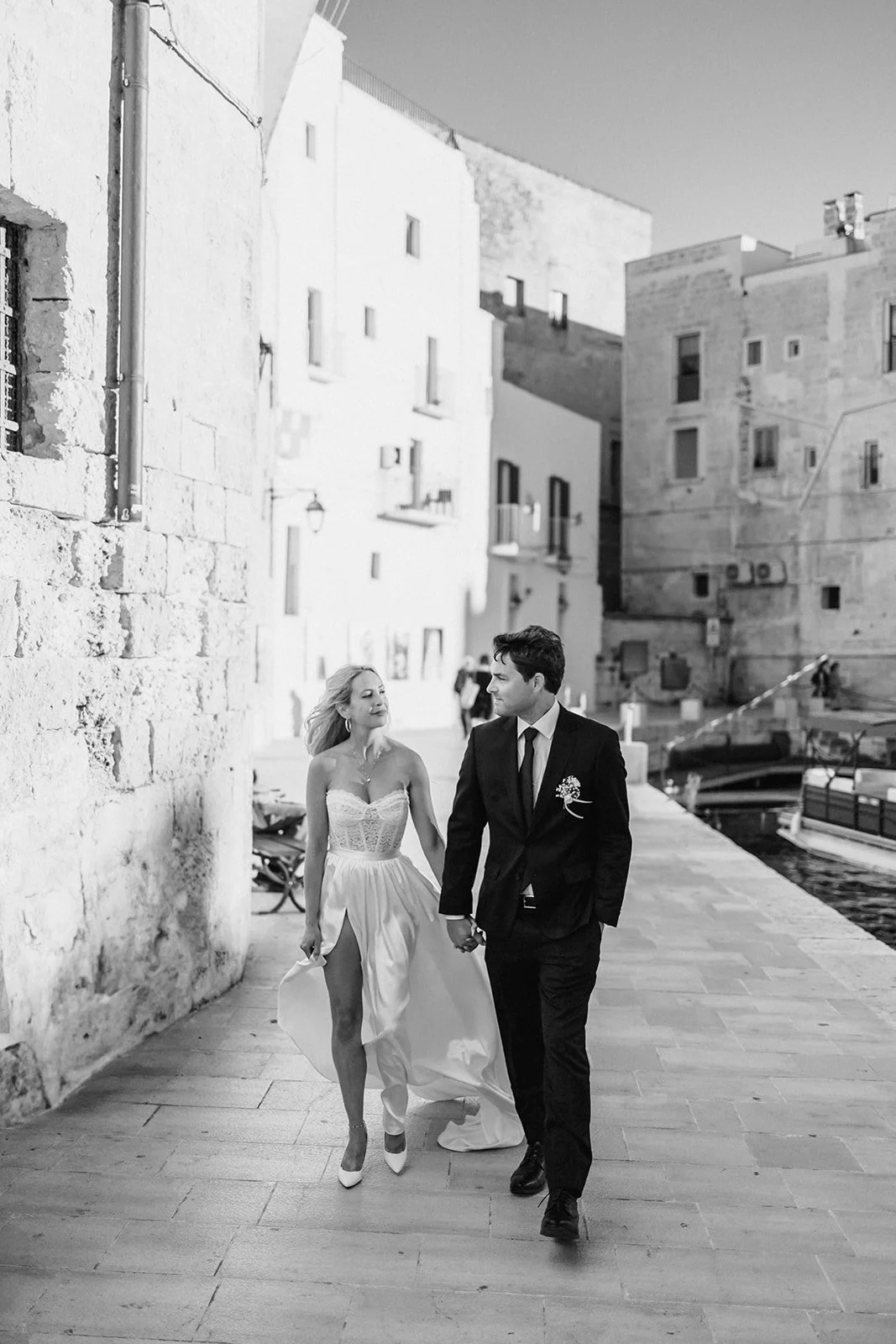 A black and white photo of a couple walking along a harbor, the woman in a wedding dress and the man in a suit, holding hands and looking at each other.