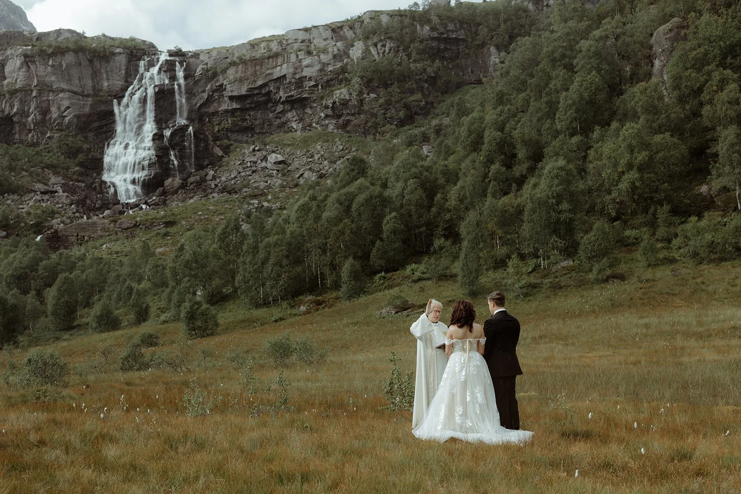 A wedding ceremony taking place outdoors in a grassy field with a waterfall and lush forest in the background.