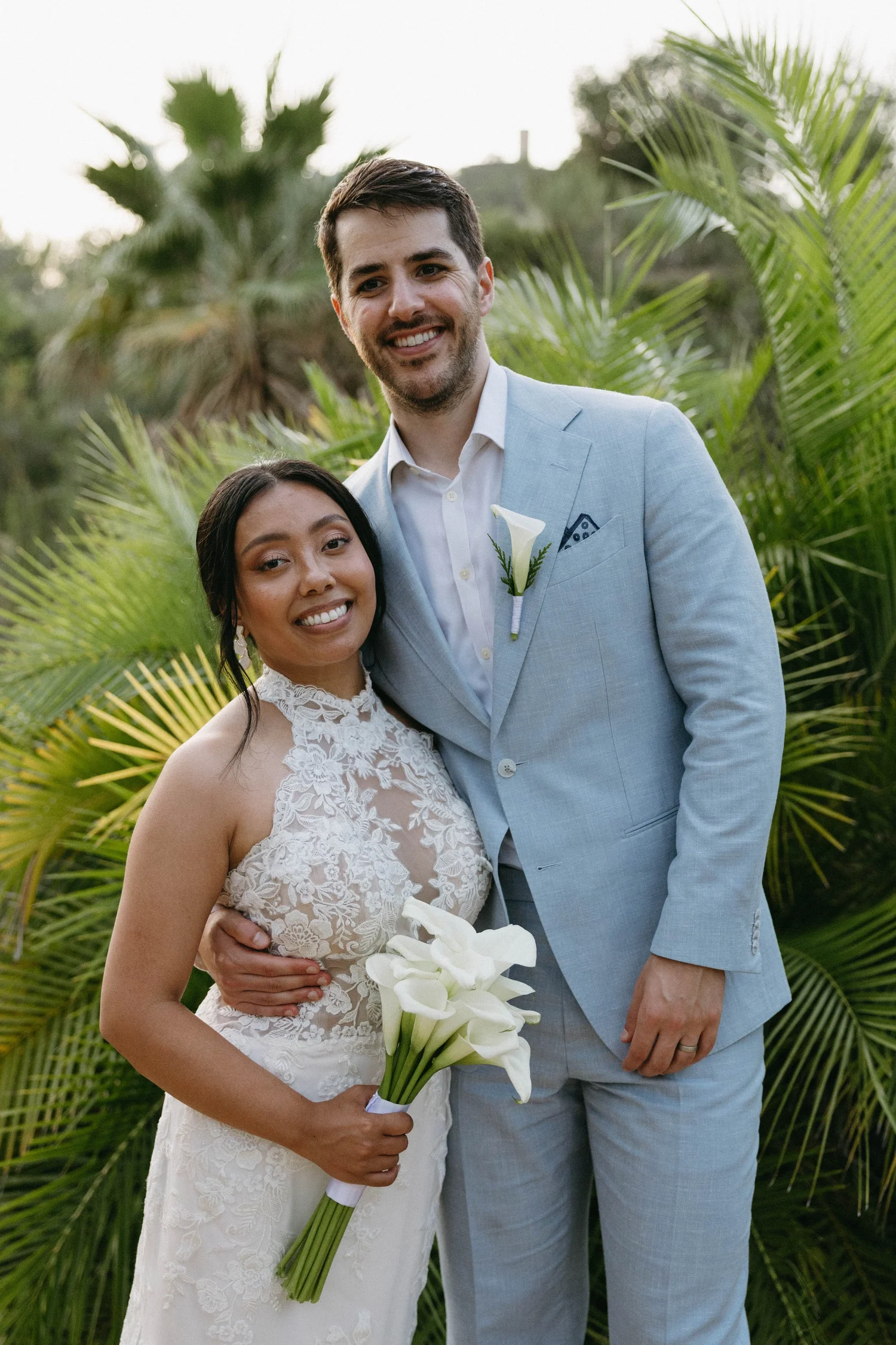 A smiling couple dressed in wedding attire, standing outdoors among lush green tropical plants, holding a bouquet of white calla lilies.