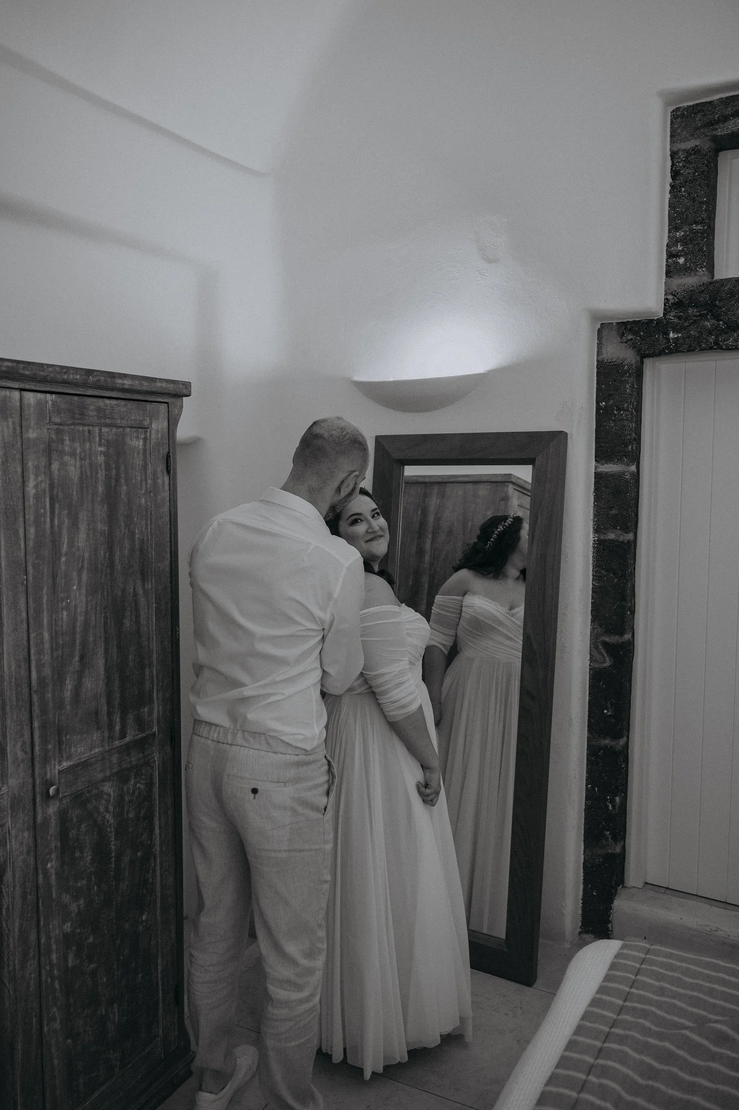 A man and a woman are looking into a full-length mirror. The man is standing behind the woman, who is dressed in a white gown, possibly a wedding dress. They are in a room with a wooden wardrobe and a stone-framed doorway.