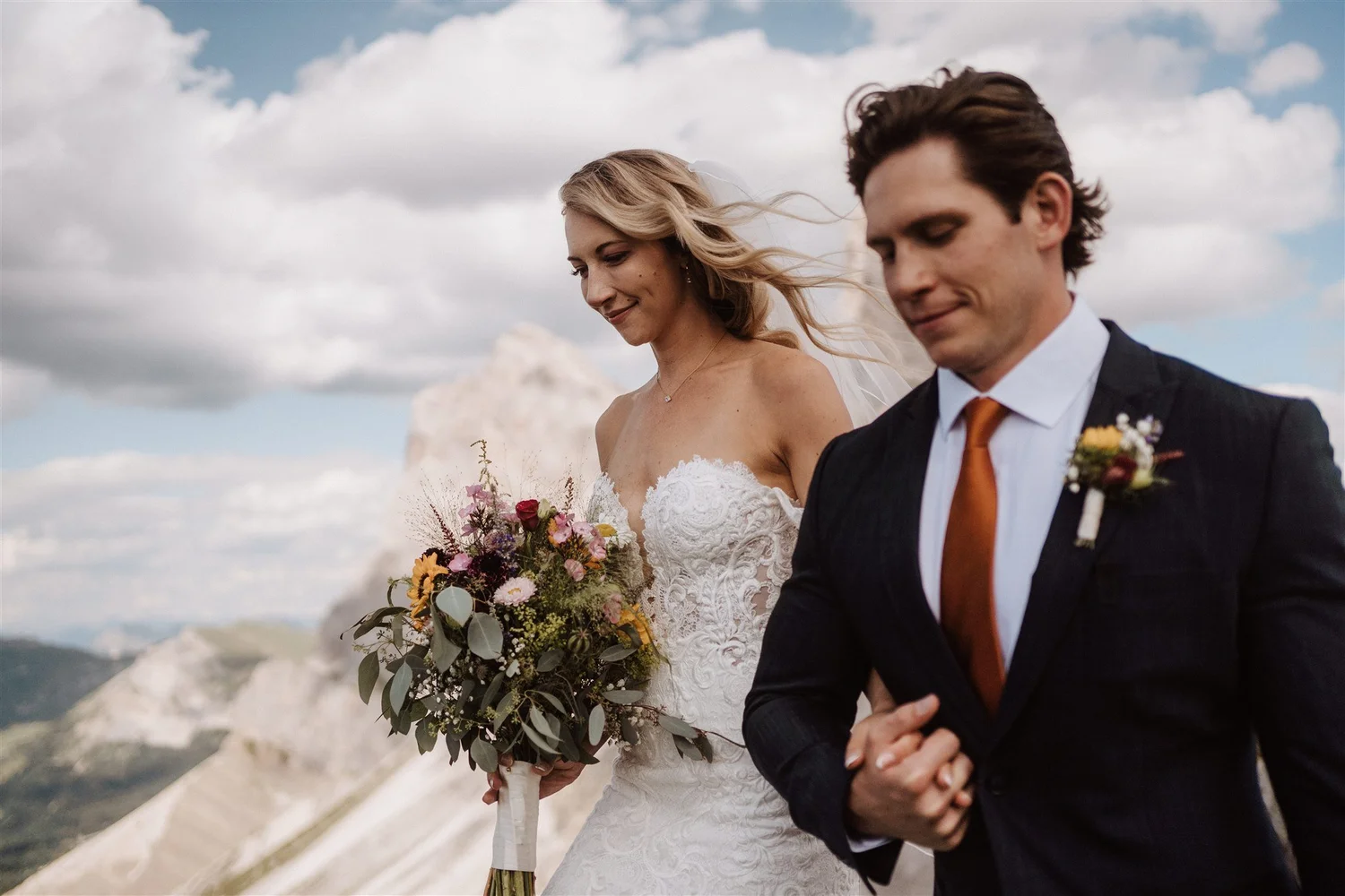 A bride and groom walking arm in arm outdoors, with the bride holding a bouquet of flowers, under a partly cloudy sky.
