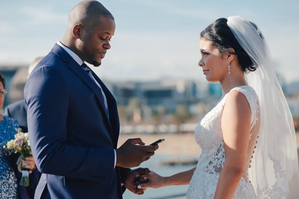 Bride and groom holding hands during their wedding ceremony outdoors, with the groom reading vows from a phone.