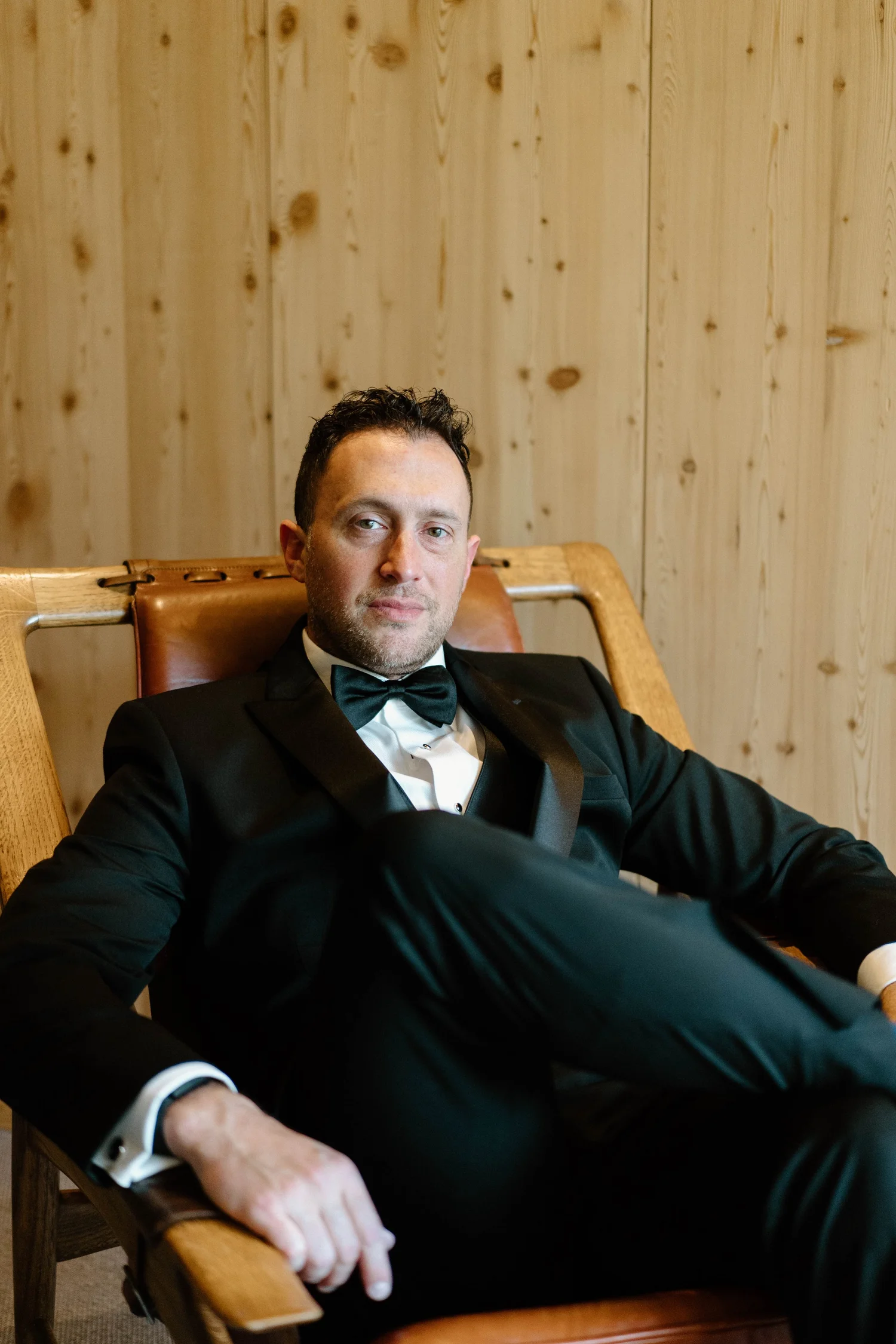 A man in a black tuxedo with a bow tie sitting on a wooden chair in front of a wood-paneled wall.