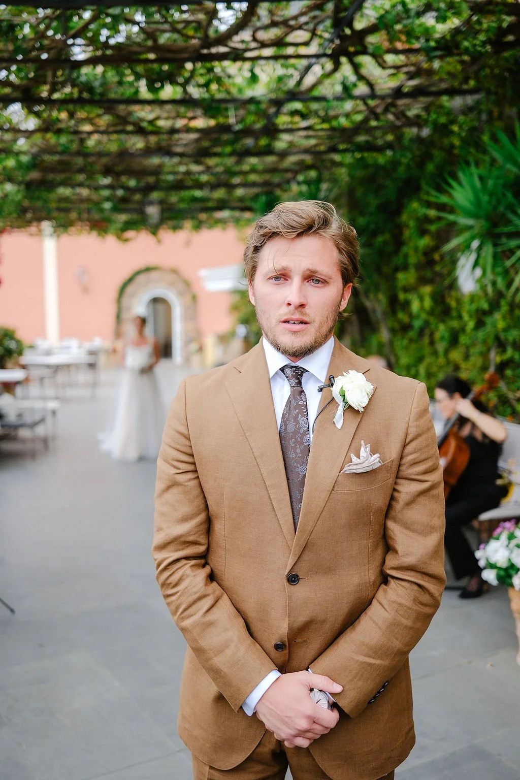 A man in a brown suit with a white boutonniere stands outdoors at a wedding ceremony, with a bride and musicians in the background.