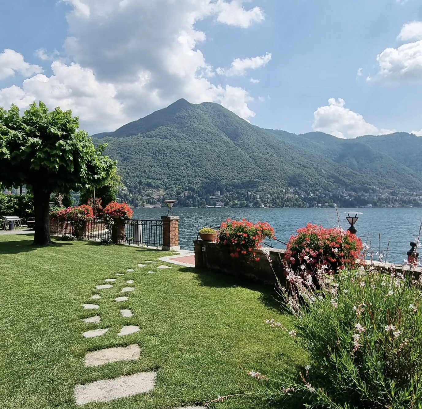 A lakeside park with a grassy area and a stepping stone pathway leading to a fence with colorful flowers, trees, and lamp posts. In the background, there is a large mountain and a body of water under a partly cloudy sky.