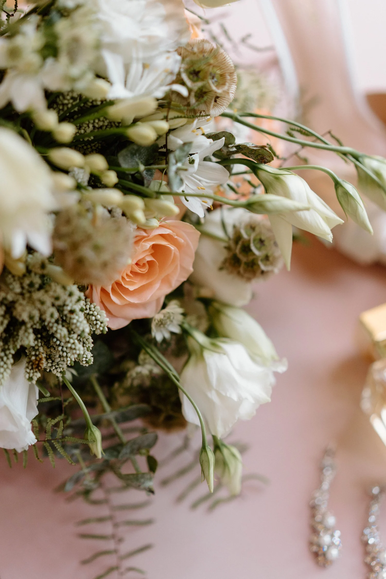 Close-up of a wedding bouquet with white and peach flowers, green foliage, and pearl embellishments.