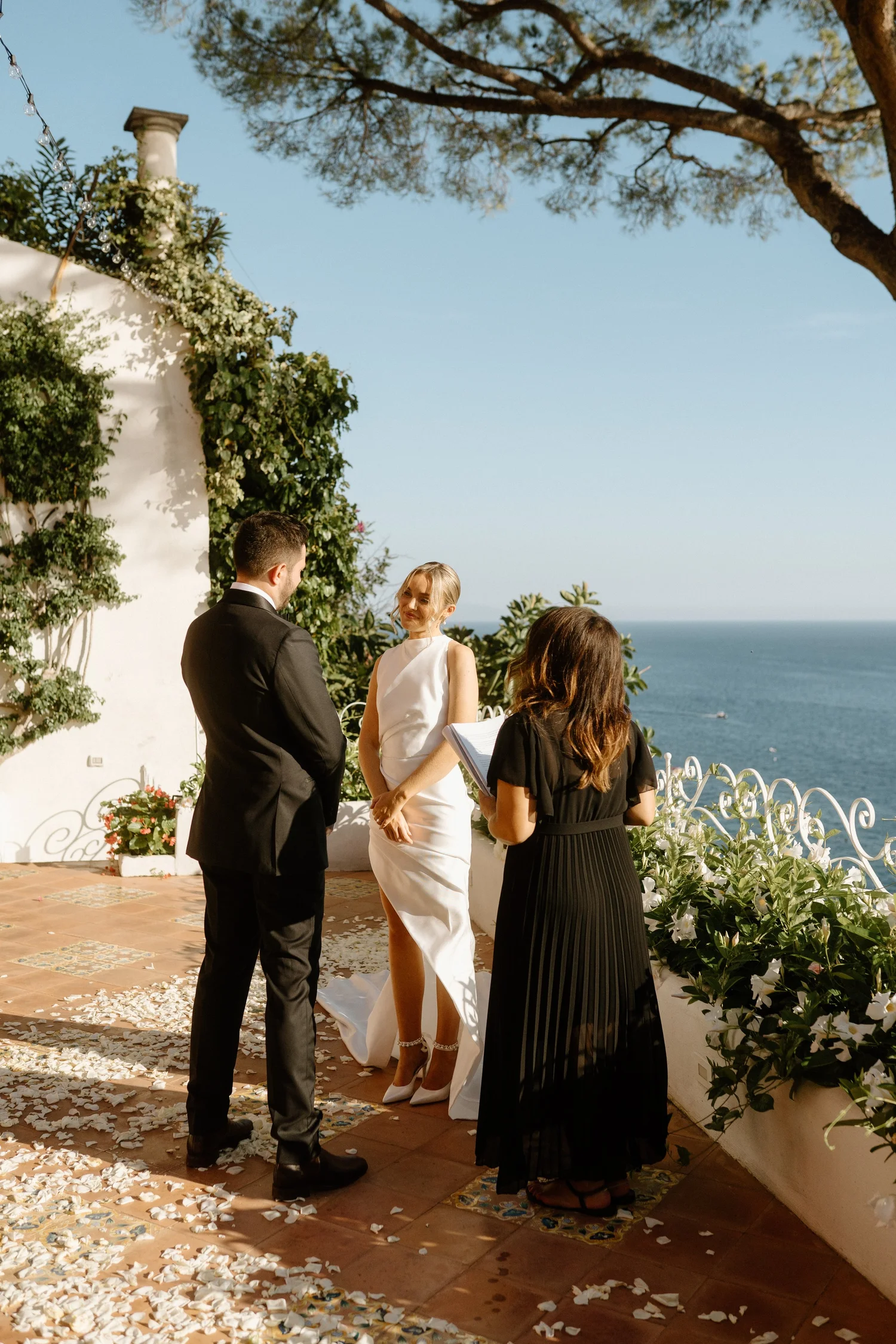 A wedding ceremony happening outdoors by the coast, with the bride and groom standing in front of an officiant. The scene is decorated with floral arrangements and white flower petals scattered on the ground, with ocean and trees in the background.