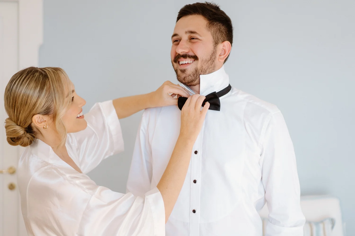 A woman is adjusting a black bowtie on a smiling man who is dressed in a white shirt with a high collar, in a light-colored room.