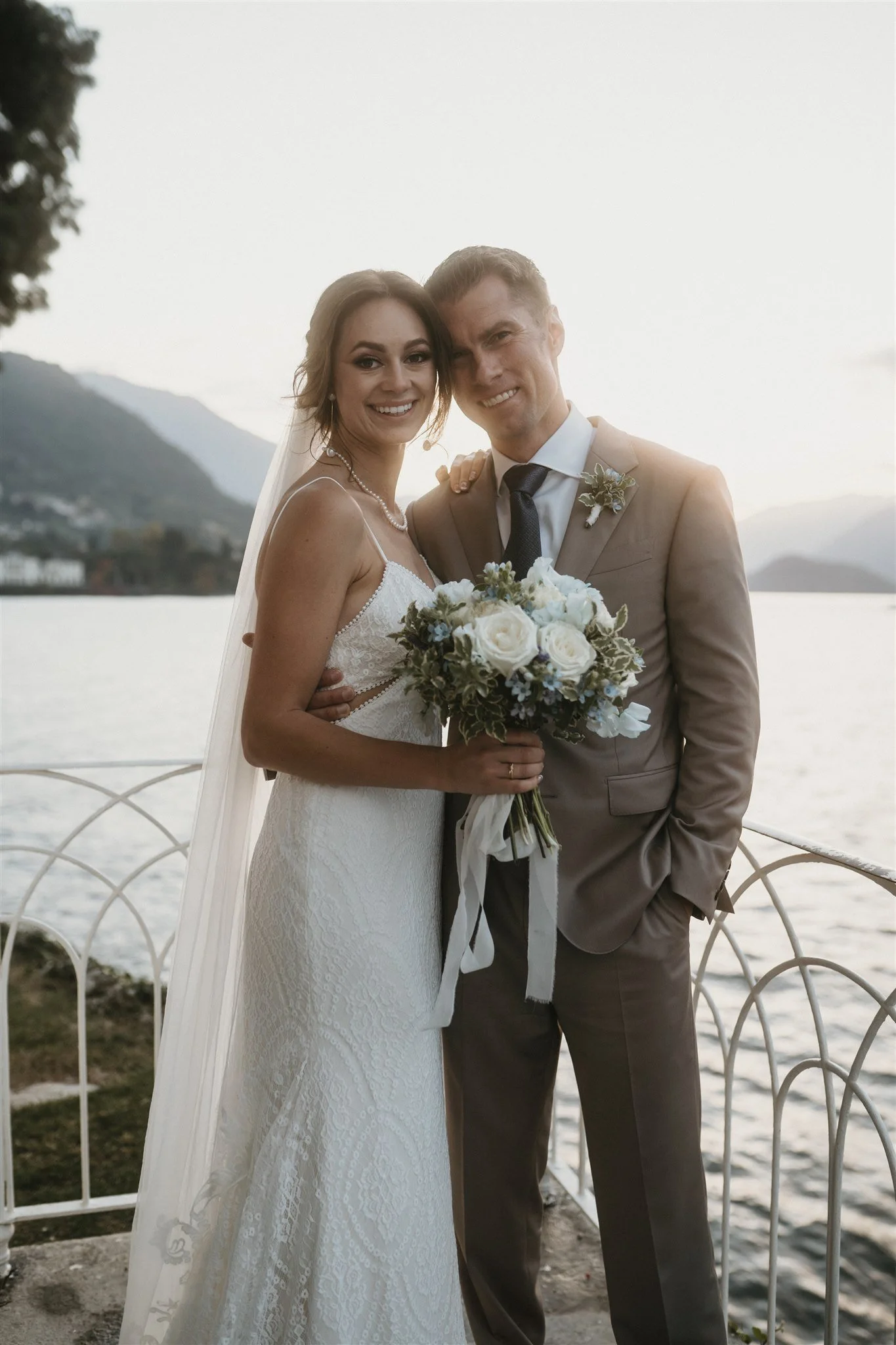 A newlywed couple embracing by a body of water, with mountains in the background, during sunset. The bride wears a white lace wedding gown and veil, holding a bouquet of white roses and greenery. The groom wears a tan suit with a tie and boutonniere,