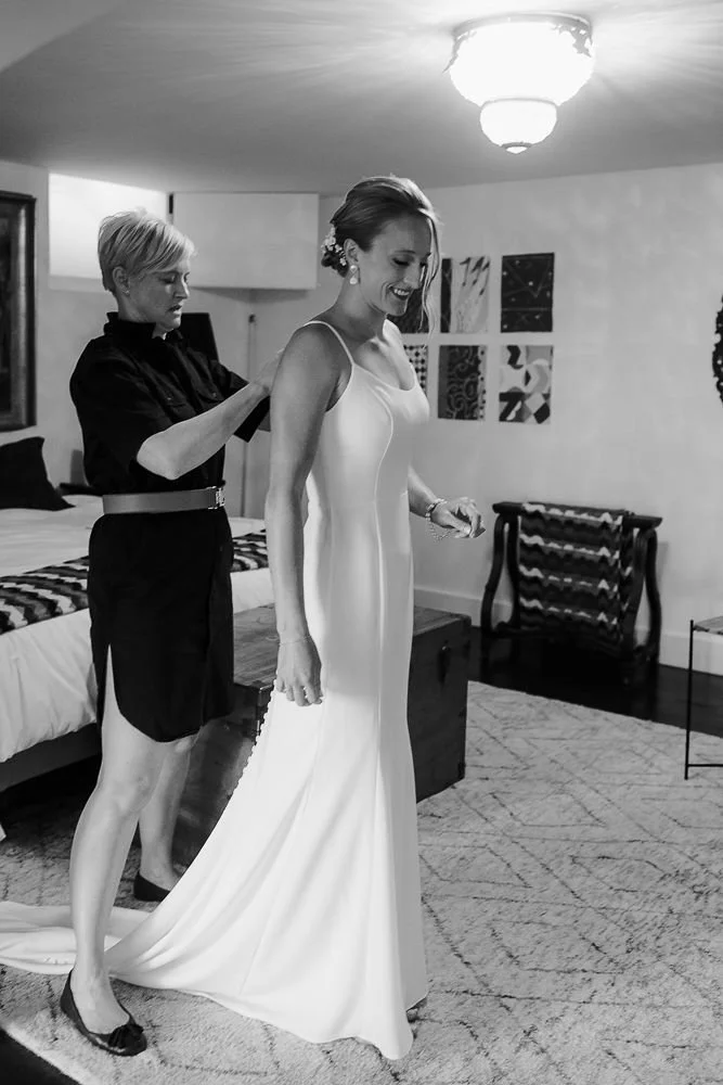 A bride in a white wedding dress is getting ready with the help of a woman in a black dress in a cozy, well-decorated room.