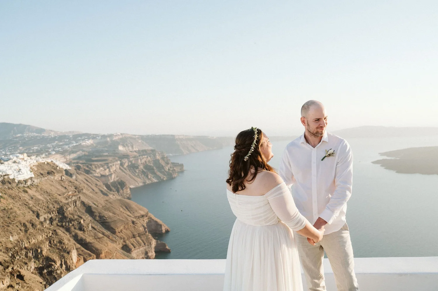 A couple dressed in white holding hands and standing on a balcony overlooking cliffs and the sea during sunset.