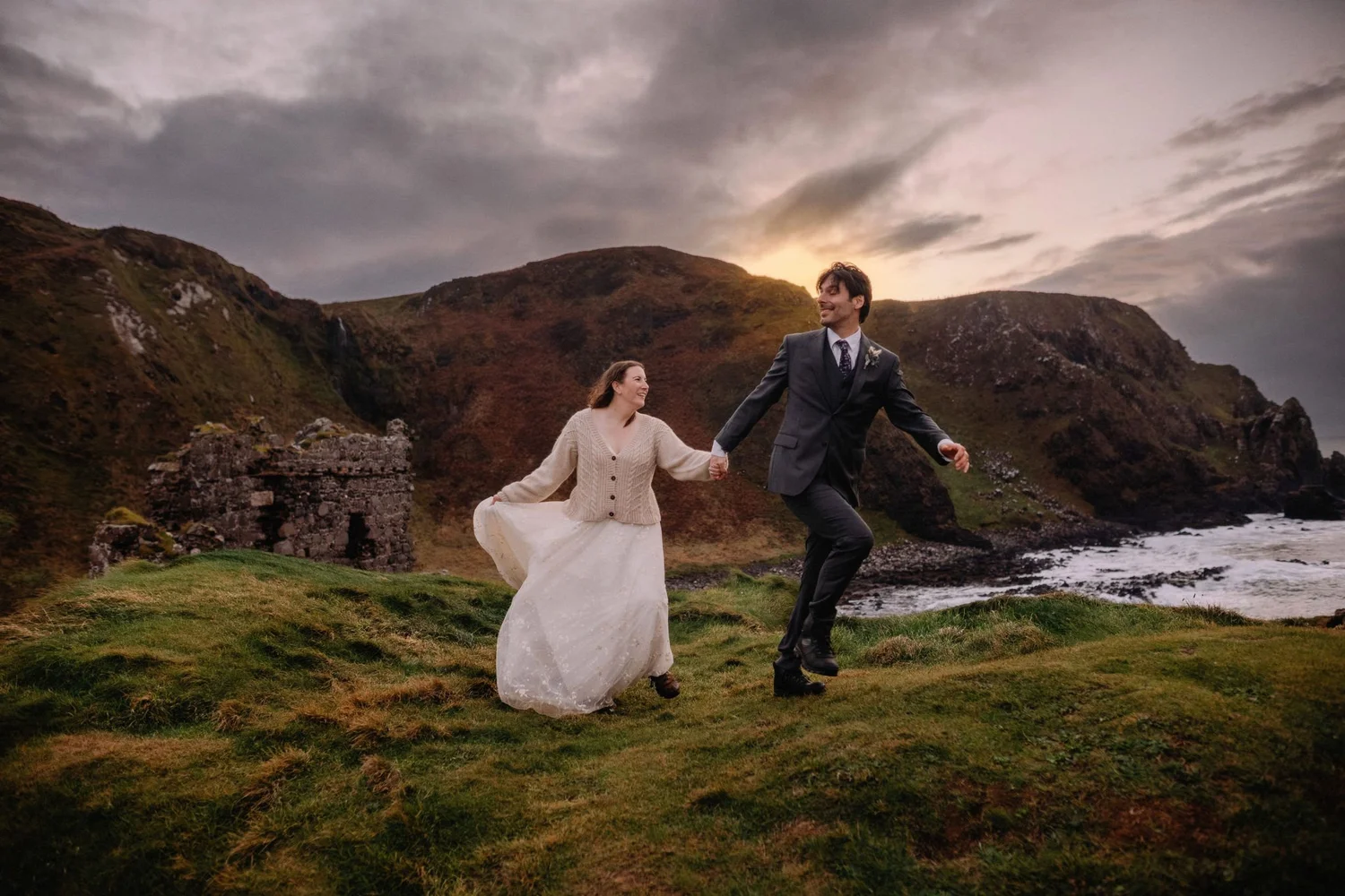 A man and woman holding hands and dancing outdoors on grassy terrain near mountains at sunset, with cloudy sky in the background.