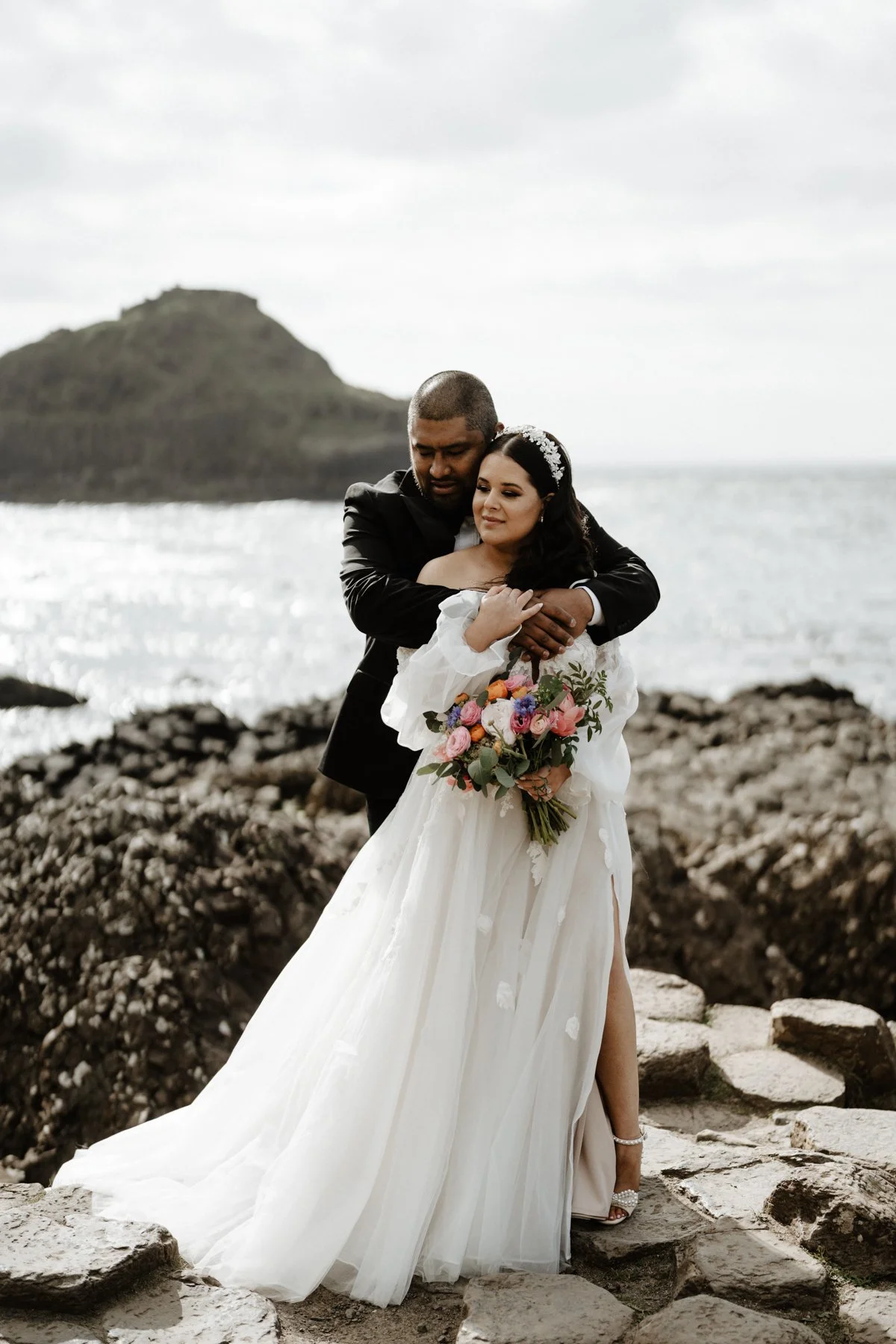 A wedding couple stands on rocky shore by the ocean, with a small island in the background. The groom in a black suit hugs the bride, who wears a white off-shoulder wedding gown and a floral headband, holding a colorful bouquet.