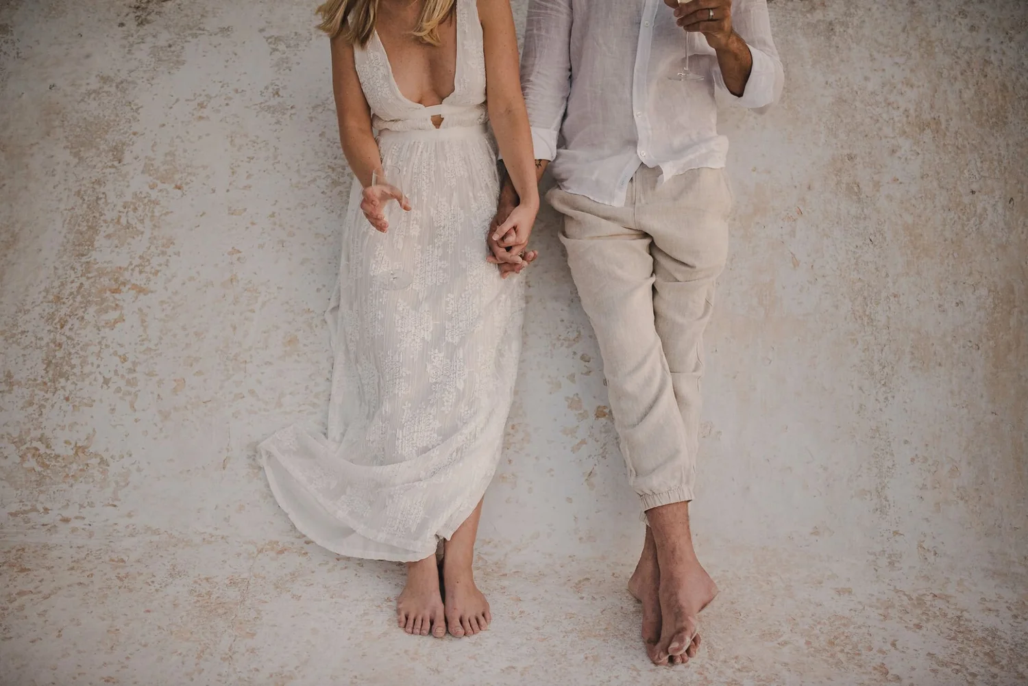 A couple standing against a textured wall, holding hands with joined fingers. The woman is wearing a white lace dress with a low neckline, and the man is dressed in light-colored linen pants and a white shirt. They are barefoot and are standing close