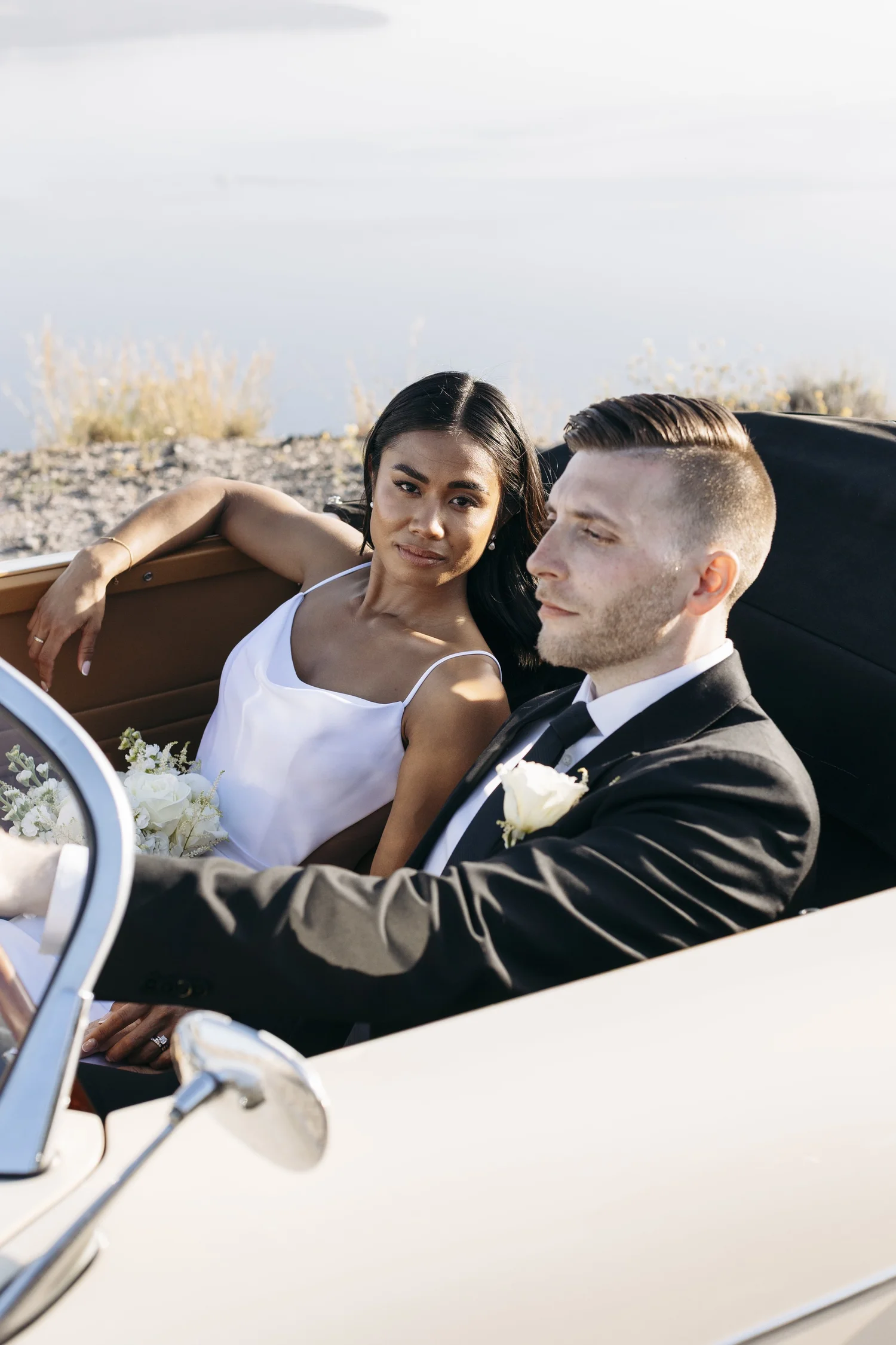 A bride and groom in a vintage convertible car during their wedding, with the bride in a white dress and the groom in a tuxedo, driving along a scenic road with water and foliage in the background.