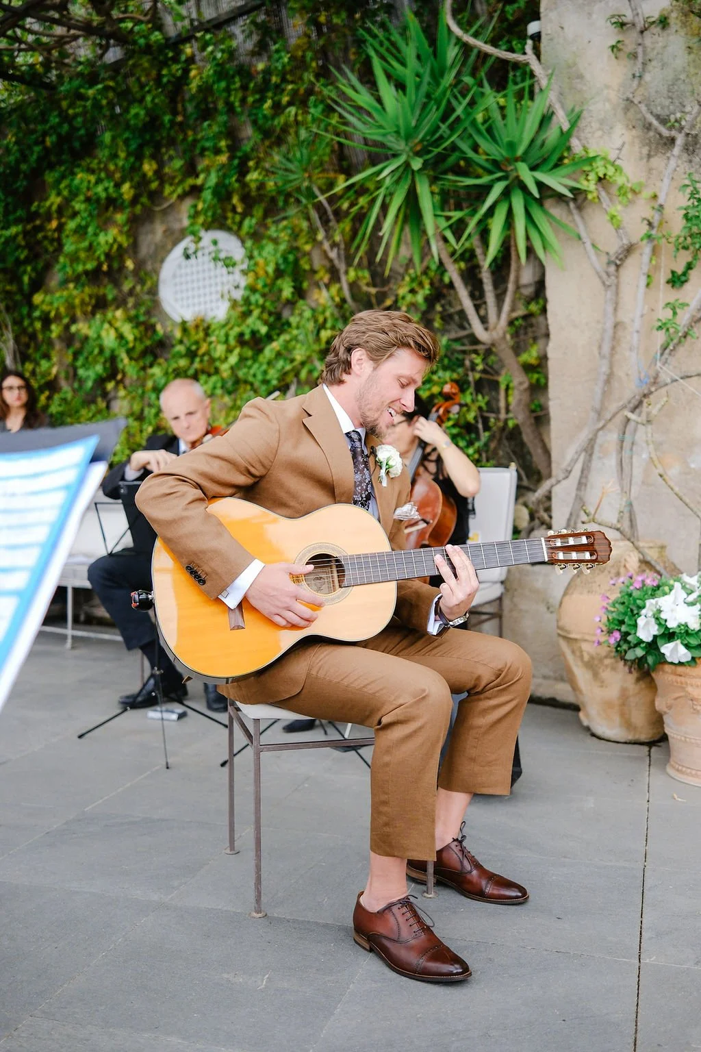 A man in a brown suit playing an acoustic guitar at an outdoor event, with a small orchestra in the background and green plants behind.