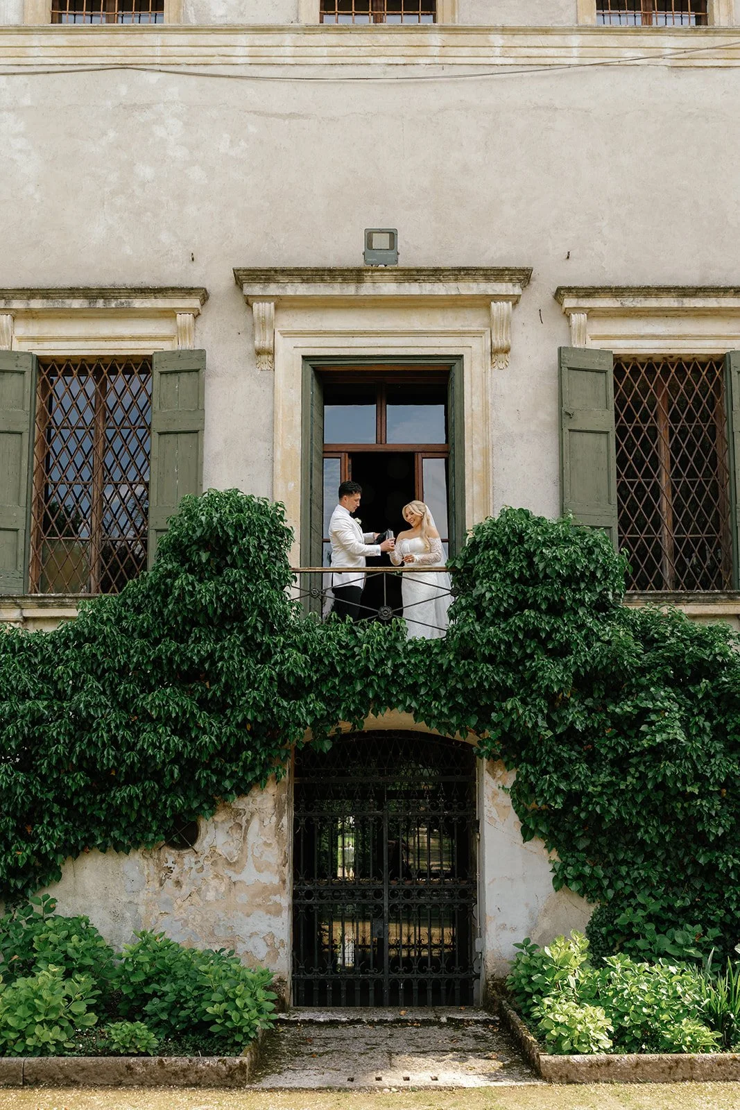 A bride and groom stand on a balcony of an old building, exchanging drinks during their wedding ceremony. The building has tall windows with wooden shutters, and green bushes surround the entrance below.