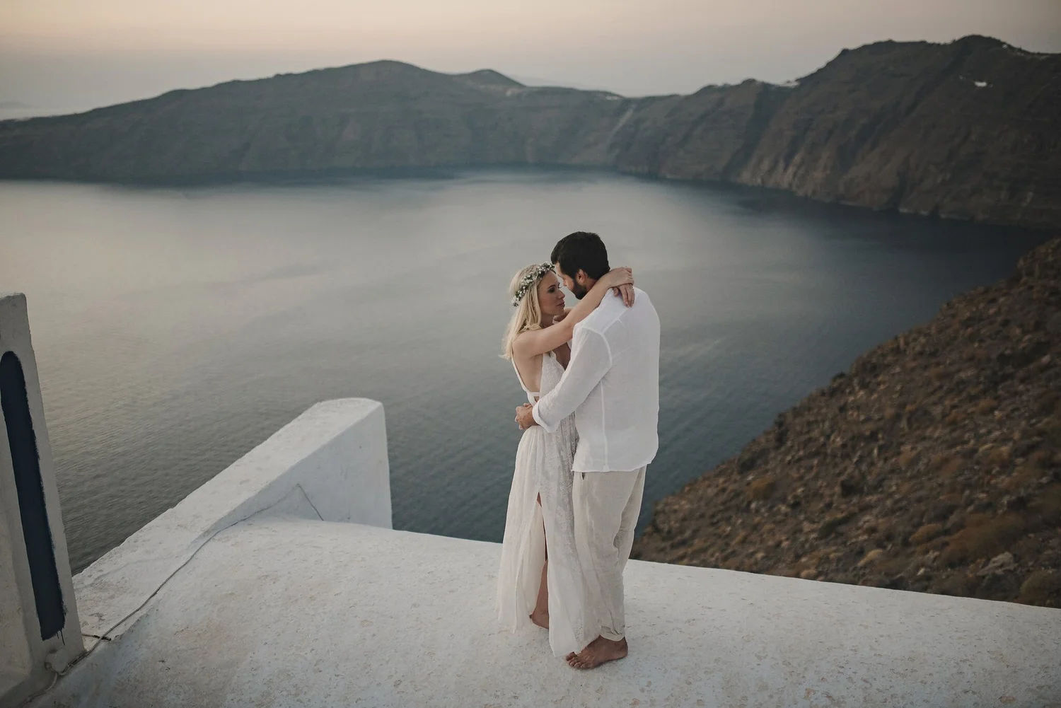 A couple embraces on a white balcony overlooking a caldera filled with water, with mountains in the background, during sunset in Santorini, Greece.