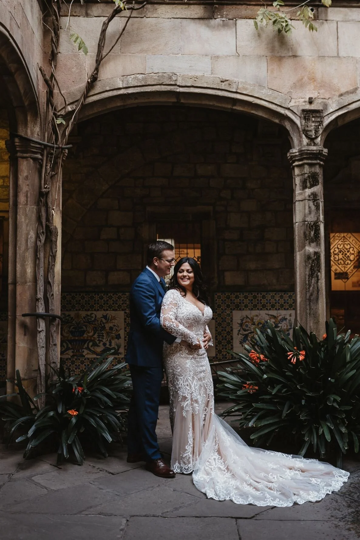 A bride and groom standing together in a courtyard with stone walls and arches, surrounded by green plants with orange flowers. The bride is wearing a white lace wedding gown with a train, and the groom is in a blue suit and glasses. They are smiling