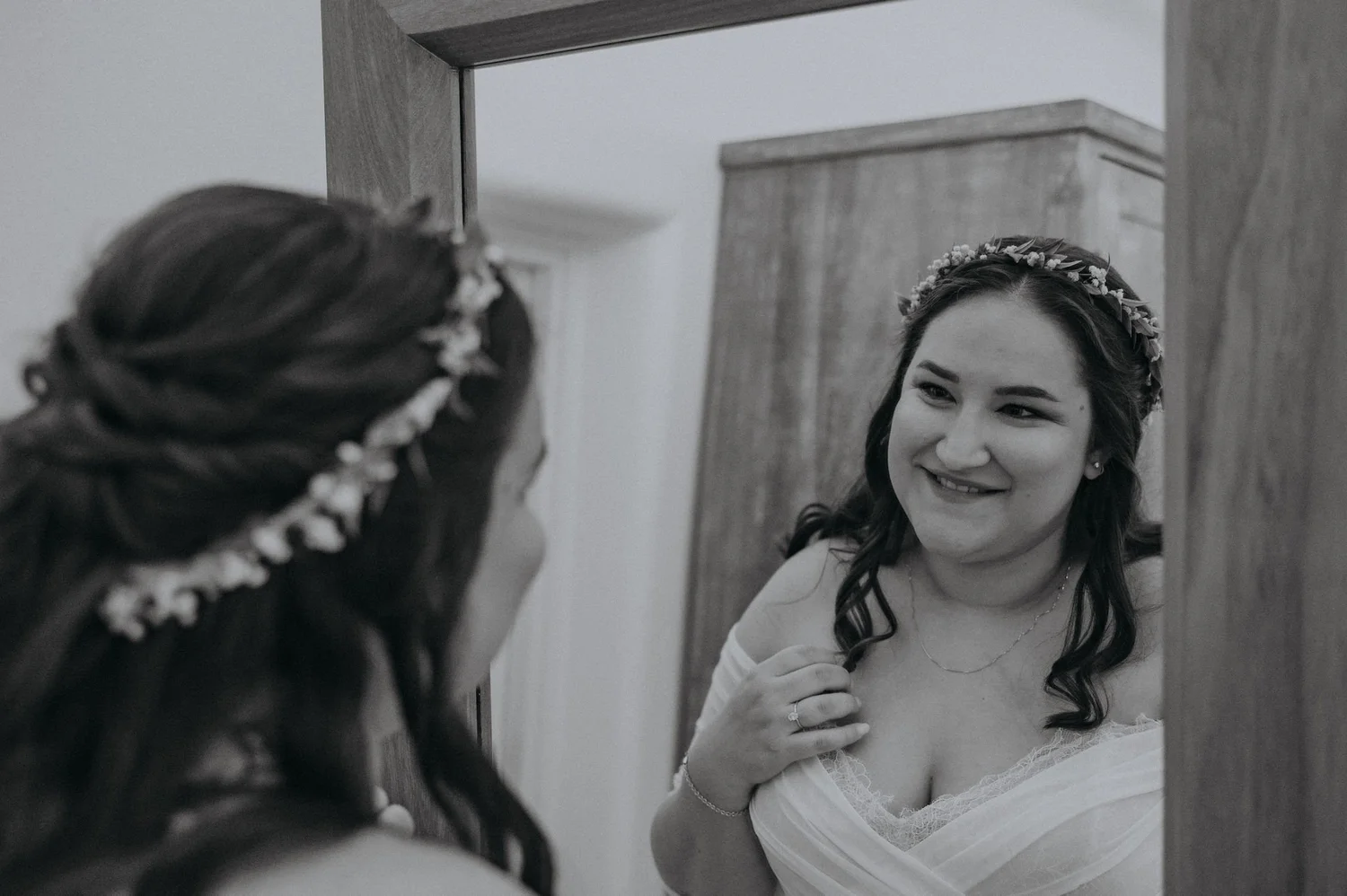 A woman with long dark hair wearing a floral headband and a white dress looks at herself in a mirror and smiles.