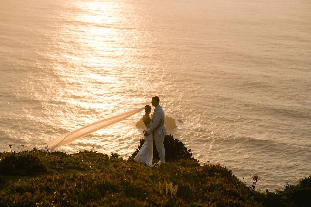A couple in wedding attire standing on a grassy cliff, embracing, with the ocean and sunset in the background.