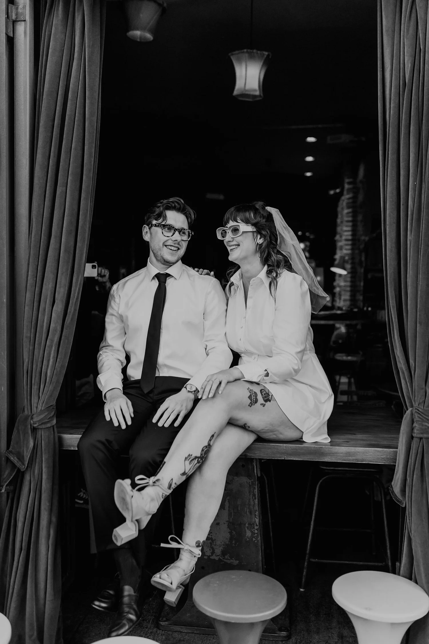 A couple sitting on a stage behind curtains, smiling at each other. The woman has tattoos on her legs and is wearing glasses, a white shirt, and a veil. The man is wearing a white shirt, black tie, and glasses. They are sitting close together and lau