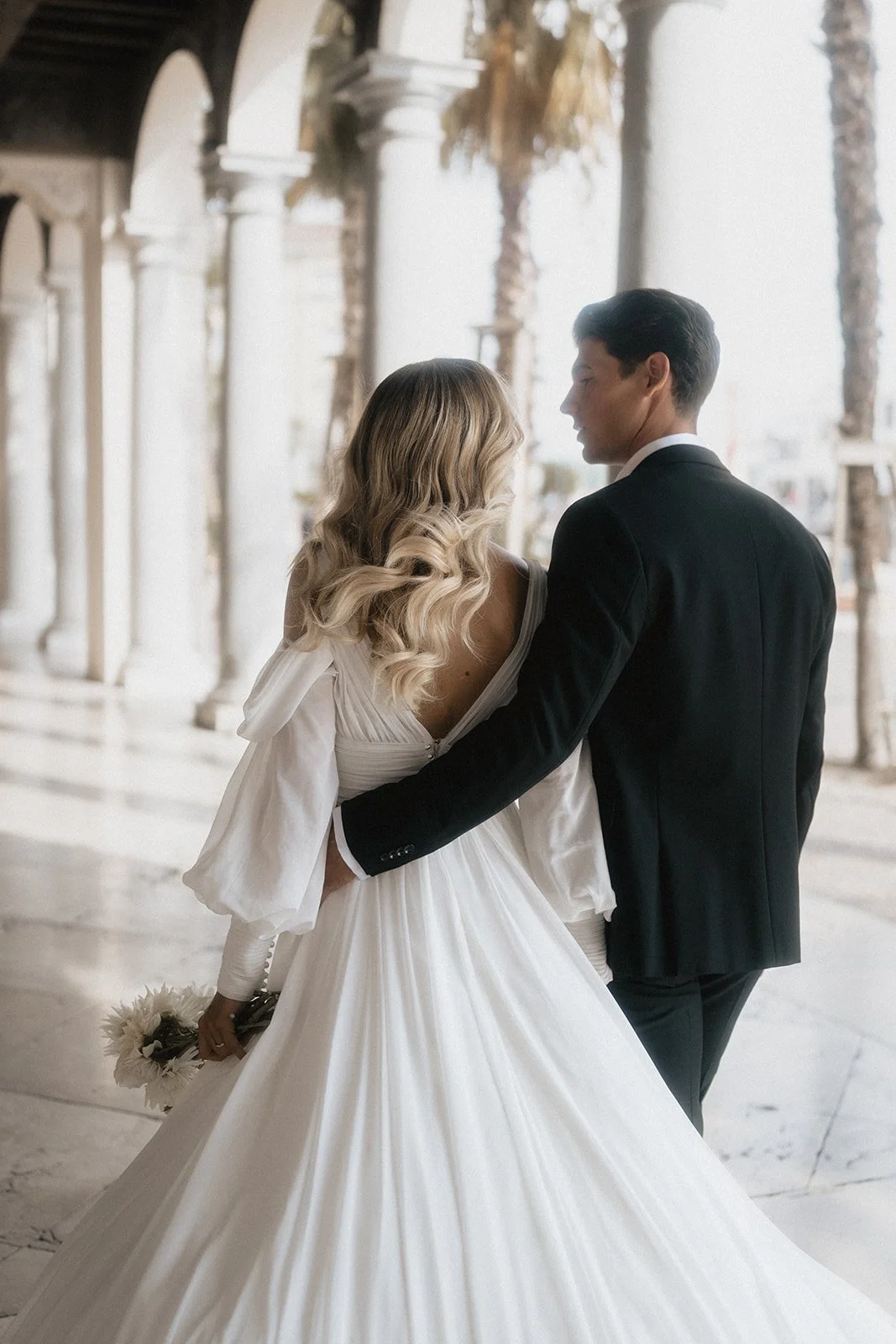 A bride and groom sharing a moment indoors, with the bride in a white gown holding flowers, and the groom in a black tuxedo, in front of a white marble building with arches and palm trees outside.