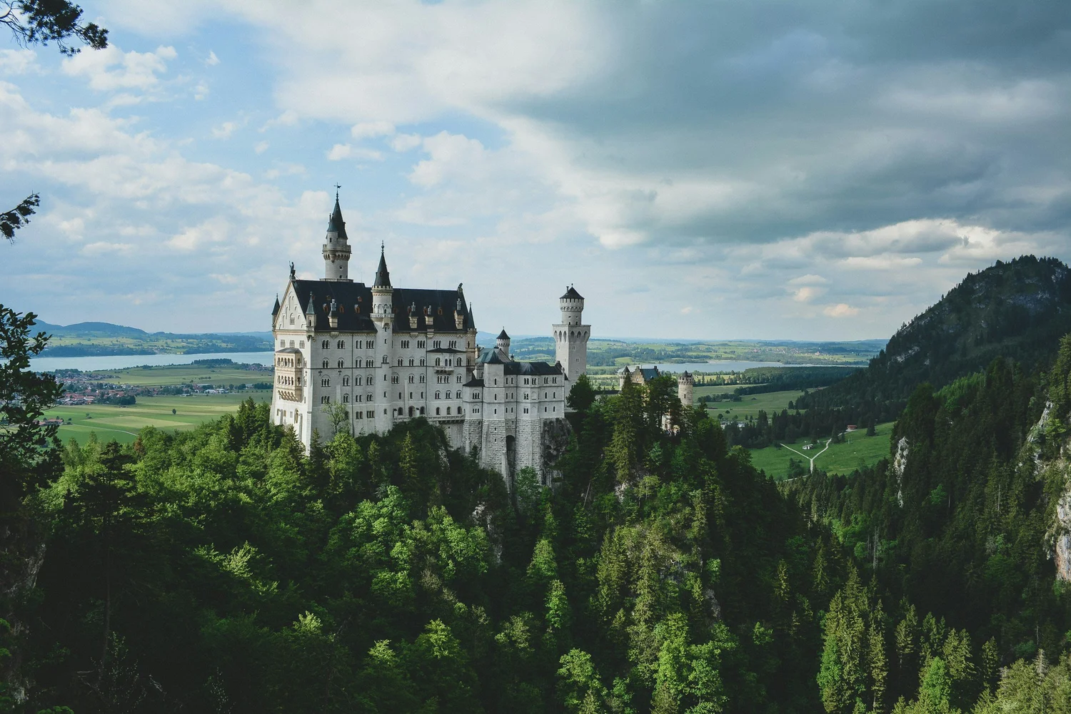 A fairy tale castle with multiple towers situated on a lush green hillside, with a river and a distant landscape of fields and hills under a cloudy sky.