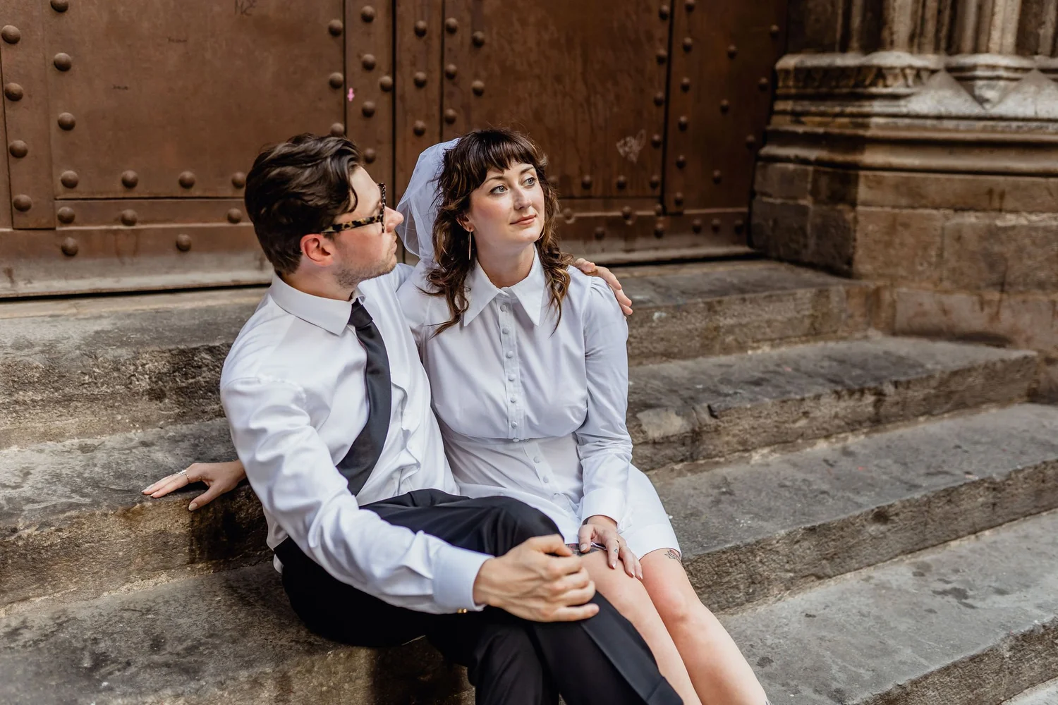 A man and woman sit on stone steps outside a historic building with large wooden doors and ornate stone columns. The woman is wearing a white shirt and veil, and the man is dressed in a white shirt and black tie. They appear to be distressed, with th