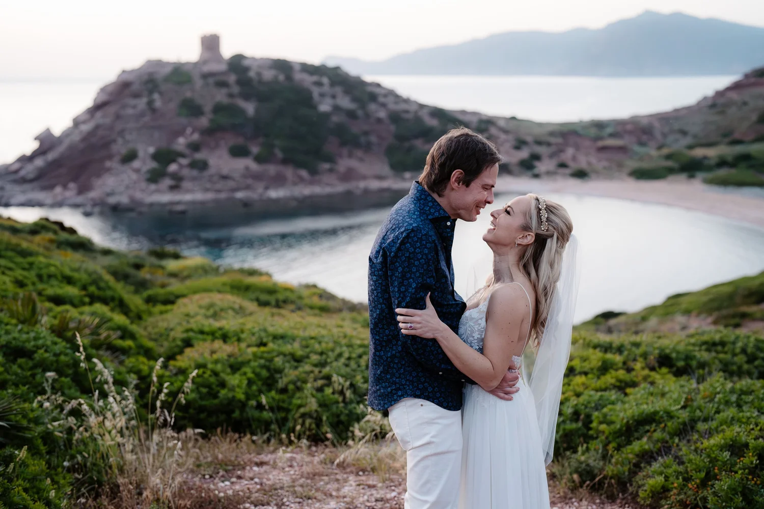 A couple dressed in wedding attire standing close together, smiling and looking into each other's eyes, outdoors near a water body with green vegetation and rocky hills in the background.