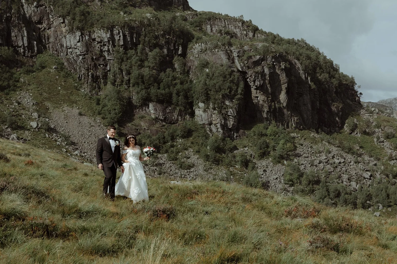 A bride and groom walking together outdoors in a grassy area with hills and rocky cliffs in the background. The bride is holding a bouquet of flowers.