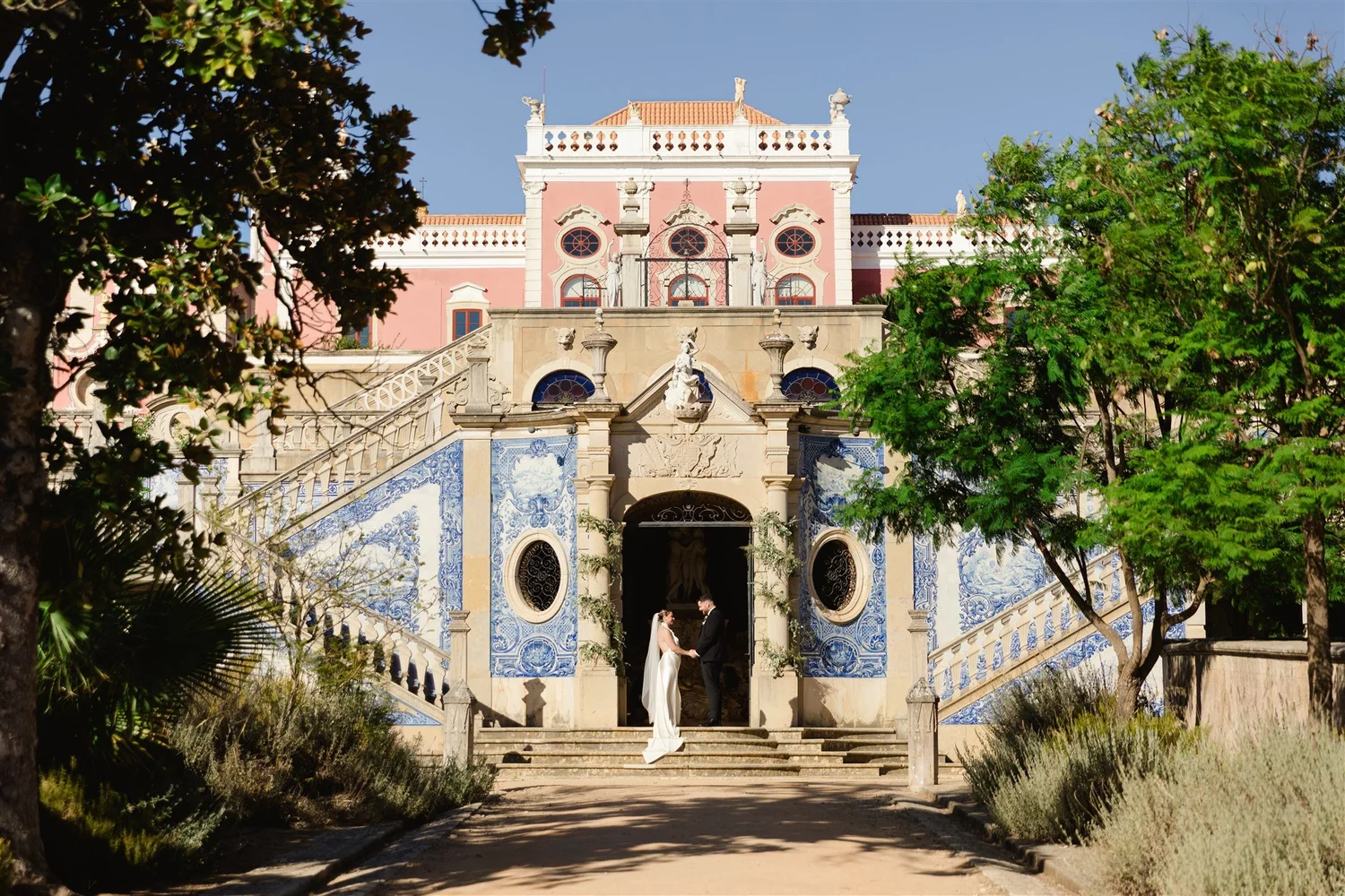 A bride and groom facing each other holding hands under an ornate entrance of a historic building with pink and white walls, surrounded by trees and greenery.