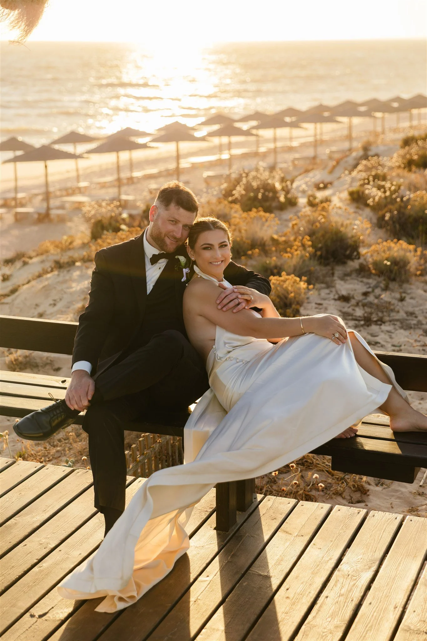 A happy couple in wedding attire sitting on a bench at the beach during sunset, with umbrellas and ocean in the background.