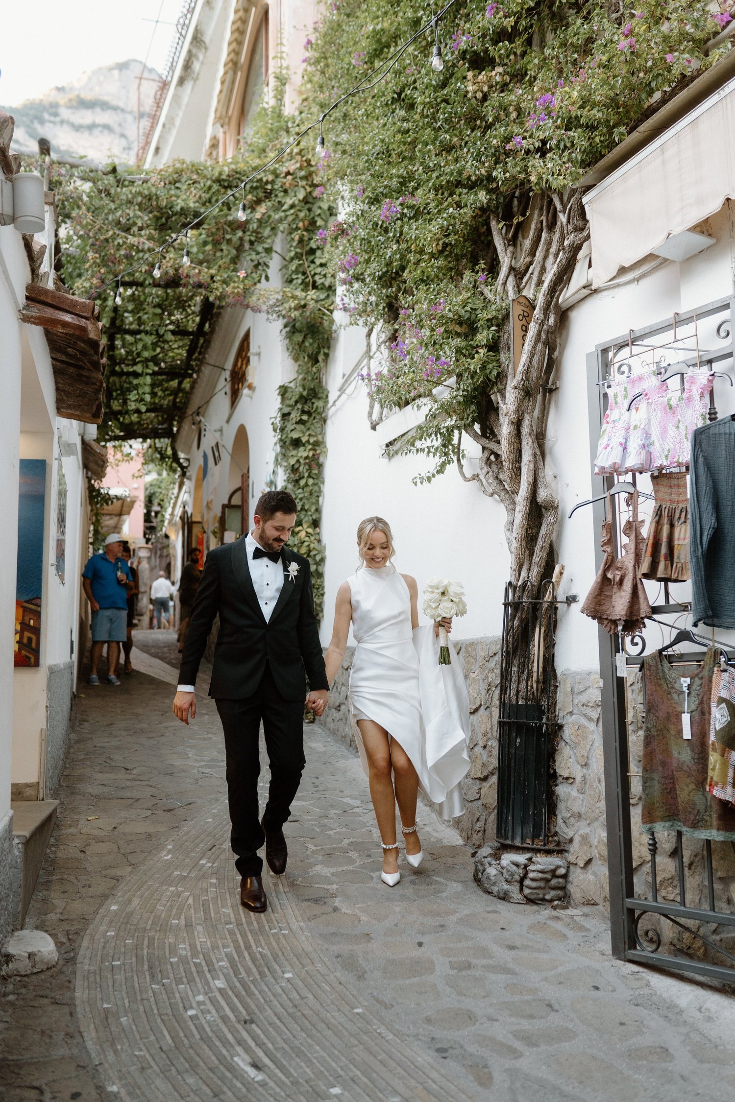 A newlywed couple walking hand in hand through a picturesque European street. The groom is wearing a black tuxedo, and the bride is wearing a white wedding dress and high heels. The street is lined with shops and decorated with string lights and purp