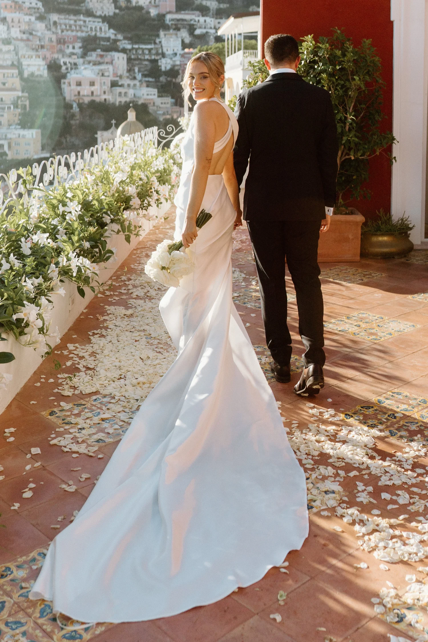 A happy bride with a white wedding dress holding a bouquet of white flowers, walking away from a groom in a black suit on an outdoor terrace decorated with flower petals, overlooking a hillside town.