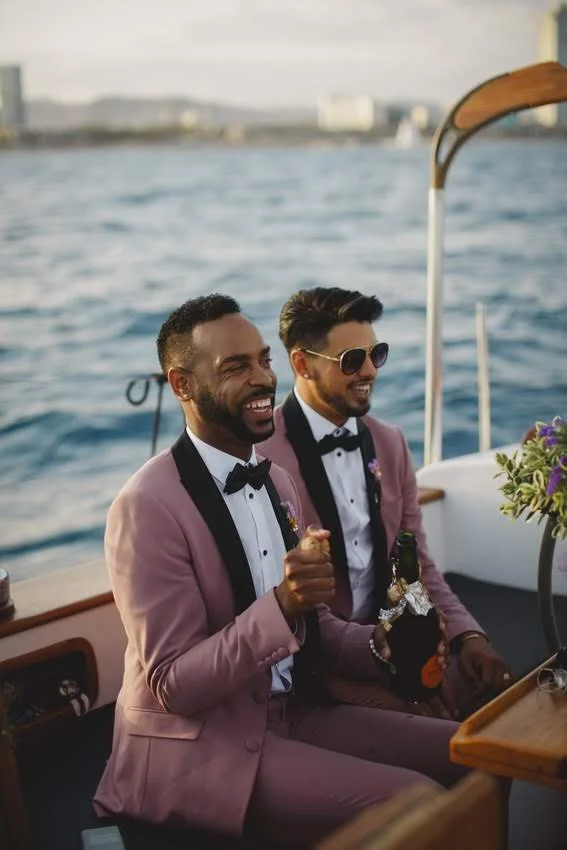 Two men dressed in pink tuxedos with black bow ties enjoying a boat ride on the water, smiling and laughing.