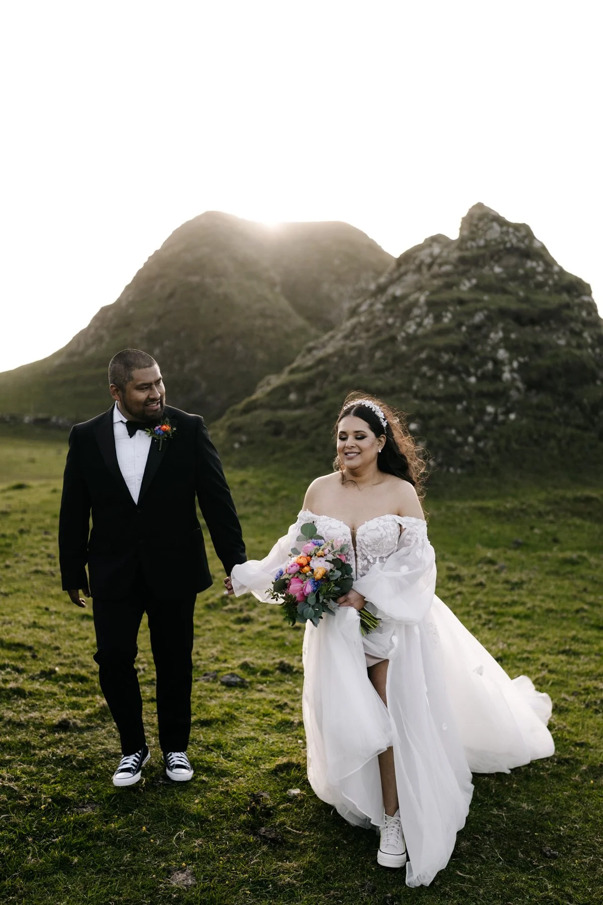 A happy couple in wedding attire holding hands in a grassy field with large, lush mountains in the background and the sun setting behind the peaks.