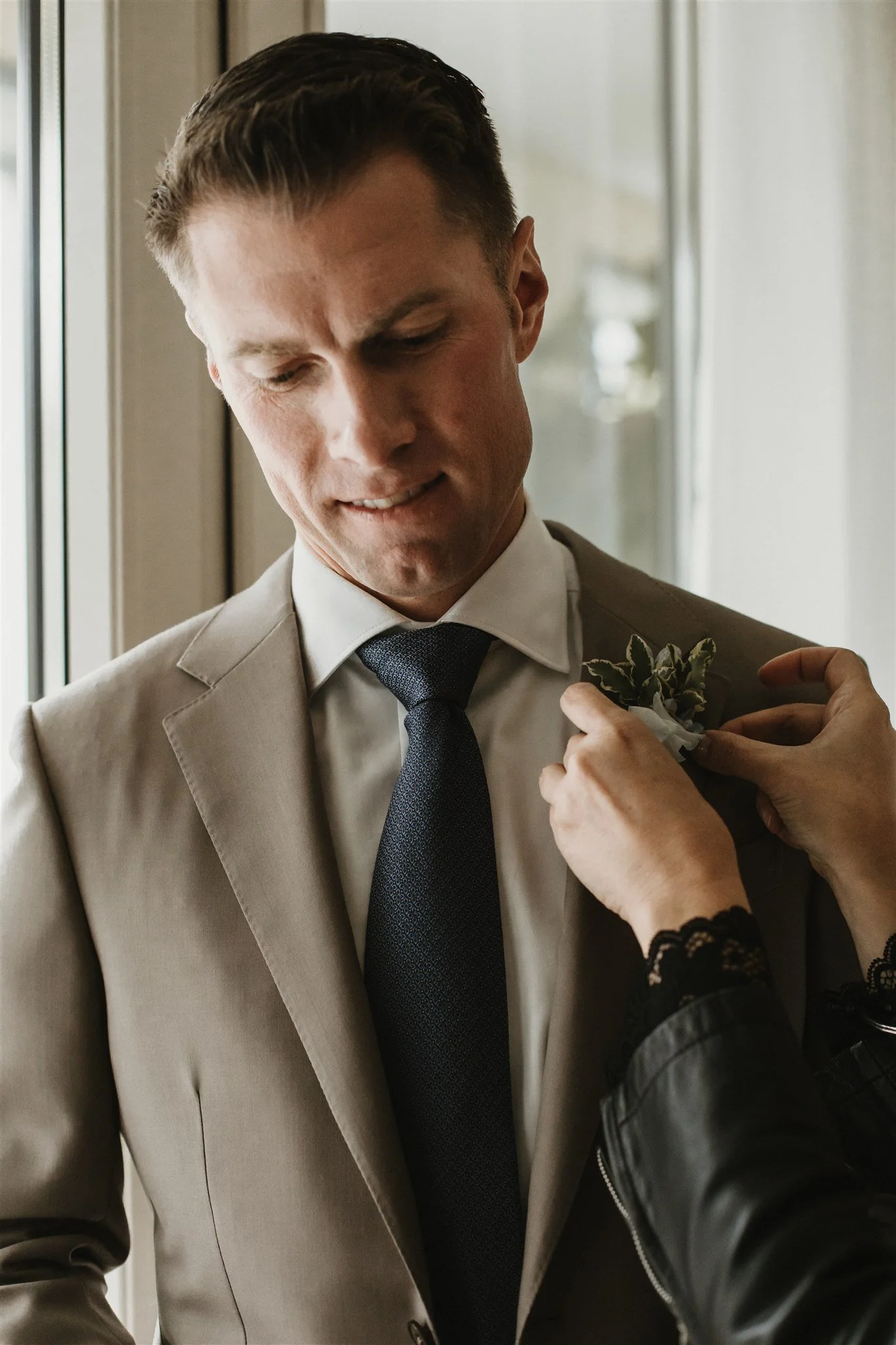 A man in a beige suit with a dark tie standing near a window, with a person in black lace sleeves pinning a boutonniere on his lapel.