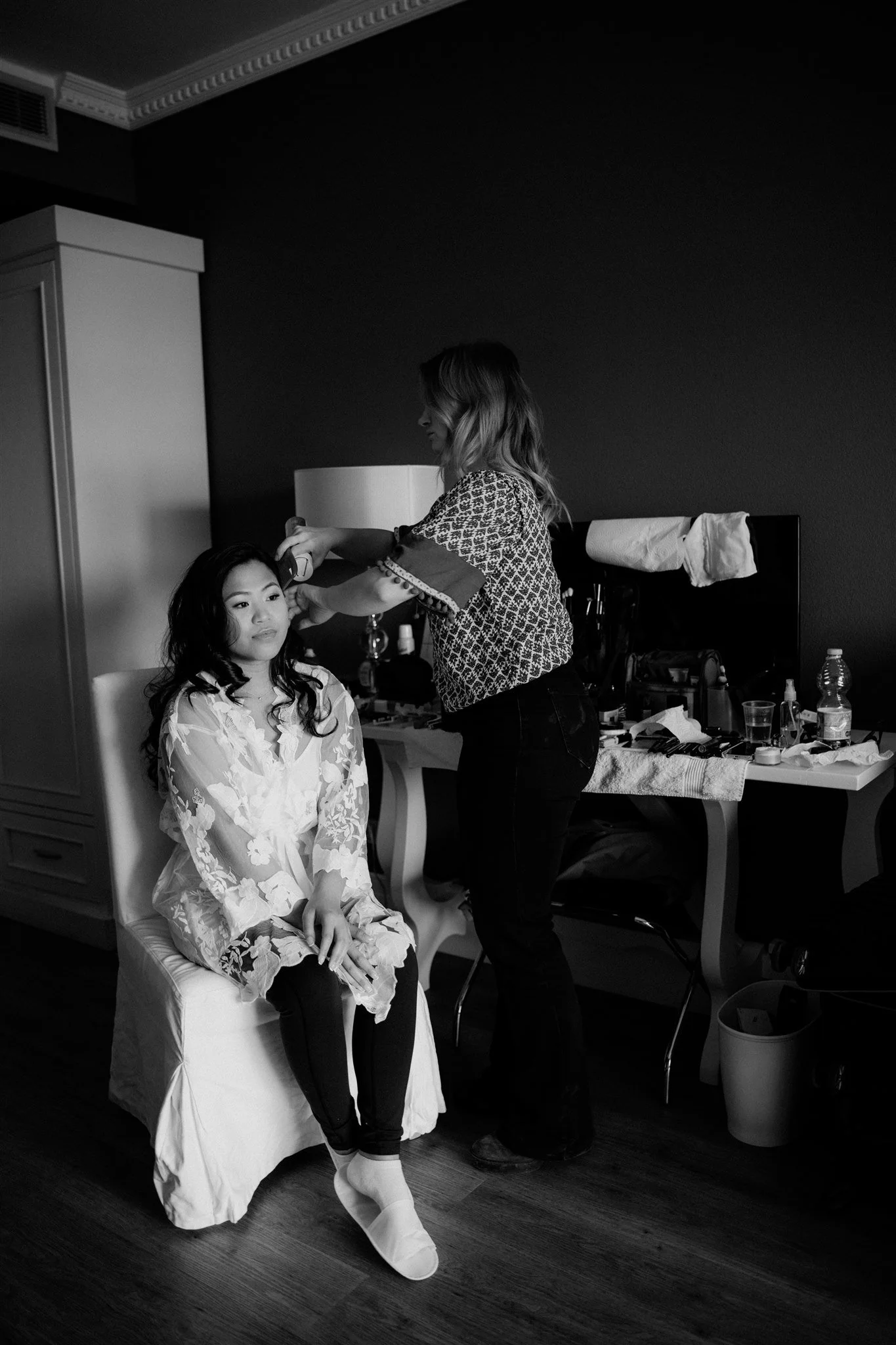 A woman sitting on a chair in a bedroom, wearing a floral robe and socks, while another woman styles her hair. The room has a table with various beauty products and tools, a lamp, and a water bottle in the background.