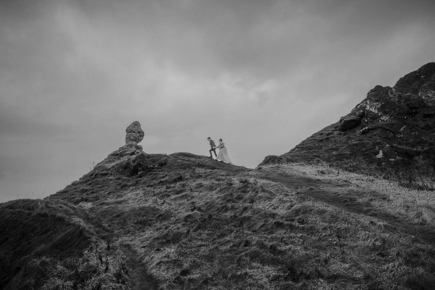 Black and white photograph of a couple holding hands and walking on a grassy hill with rocks, under a cloudy sky.