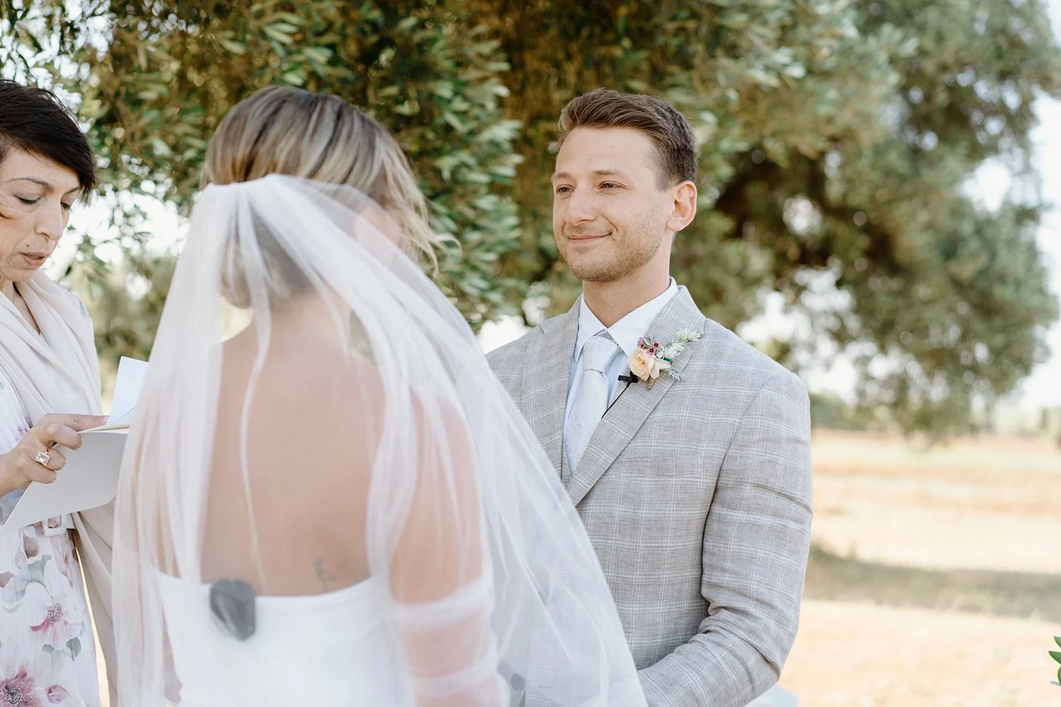 Groom with light gray suit and floral boutonniere smiling at bride during outdoor wedding ceremony under trees.