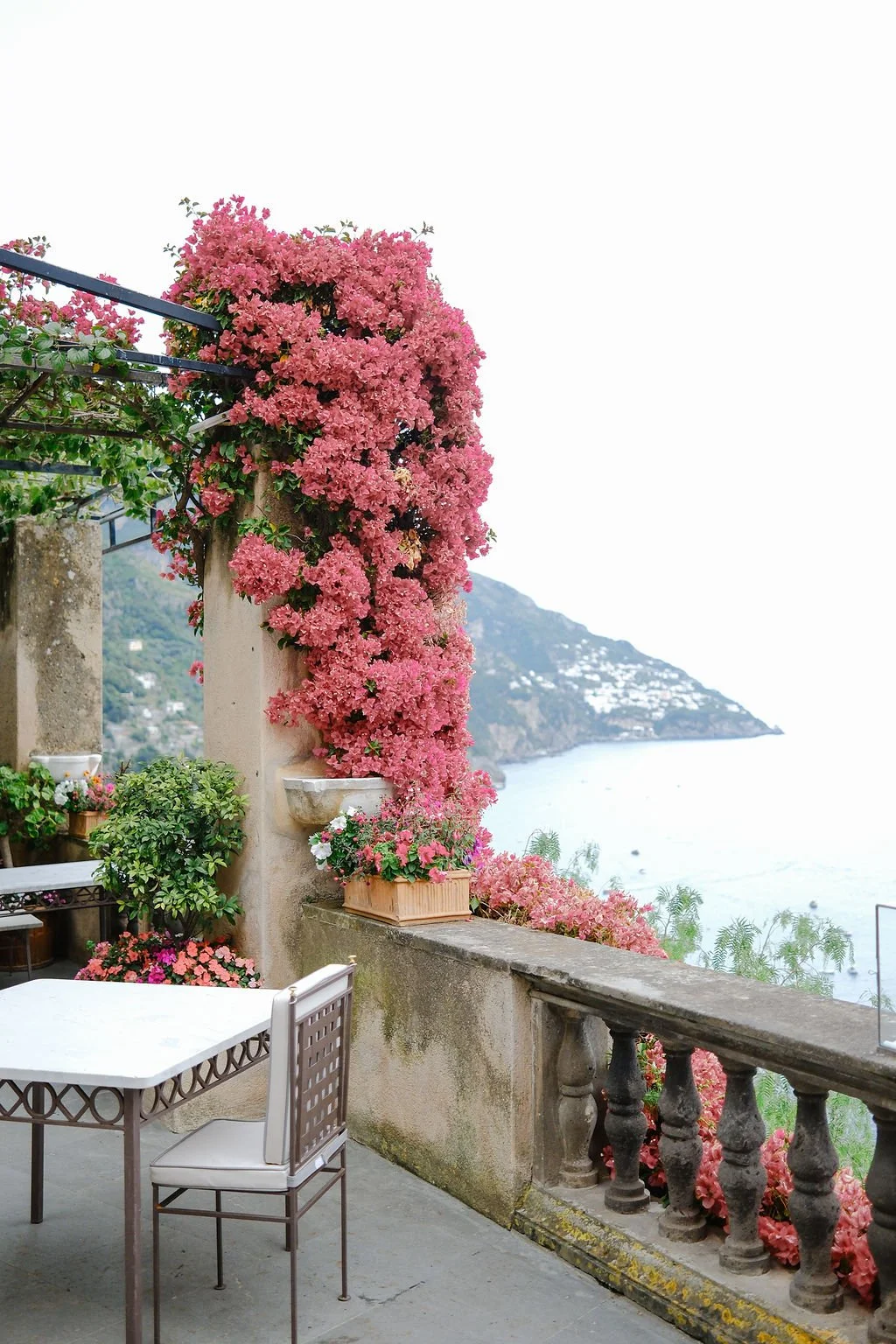 A terrace with a white table and chairs, decorated with pink flowering plants and an ocean view in the background.