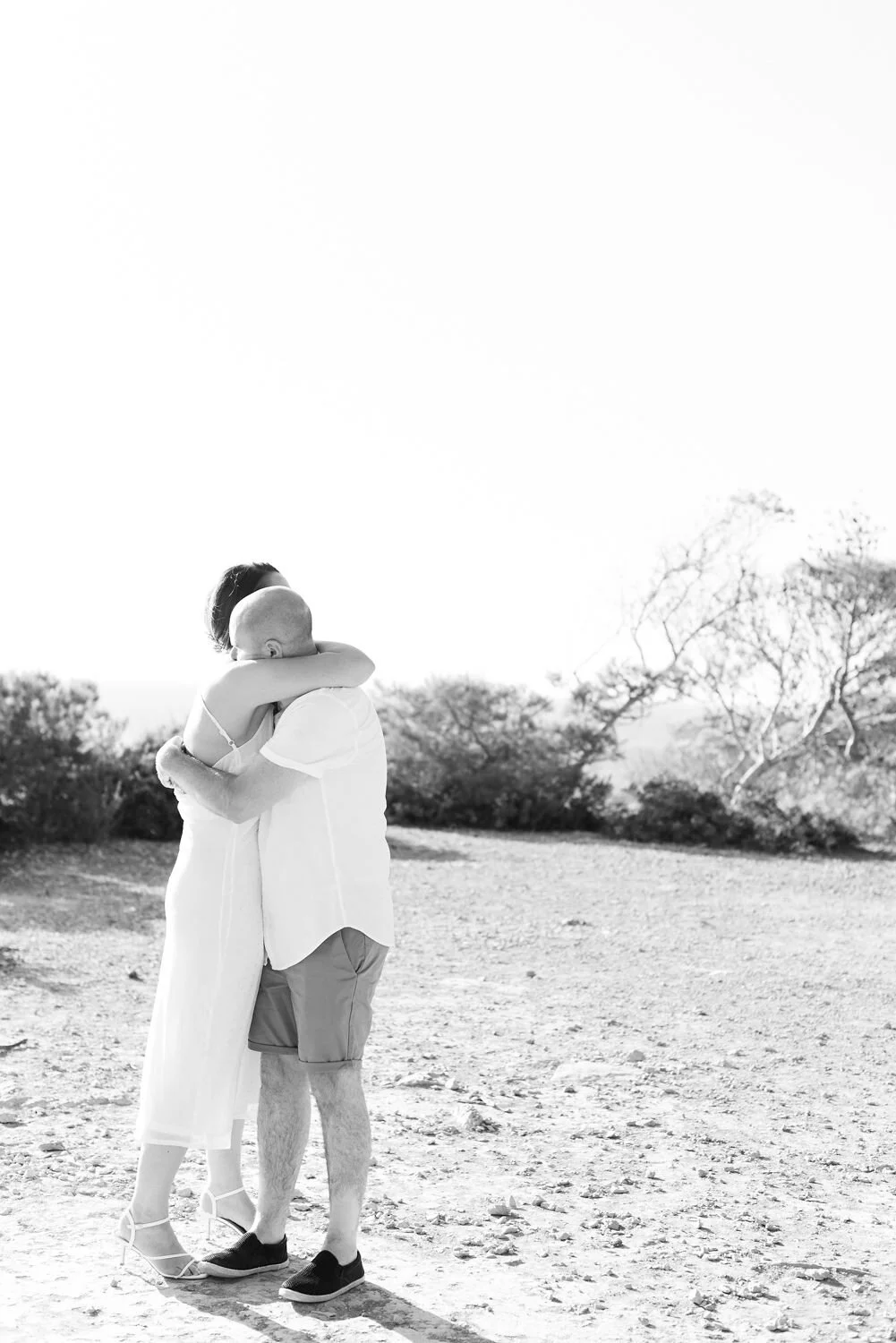 A black and white photo of two people hugging outdoors on a sunny day, with trees and an open sky in the background.